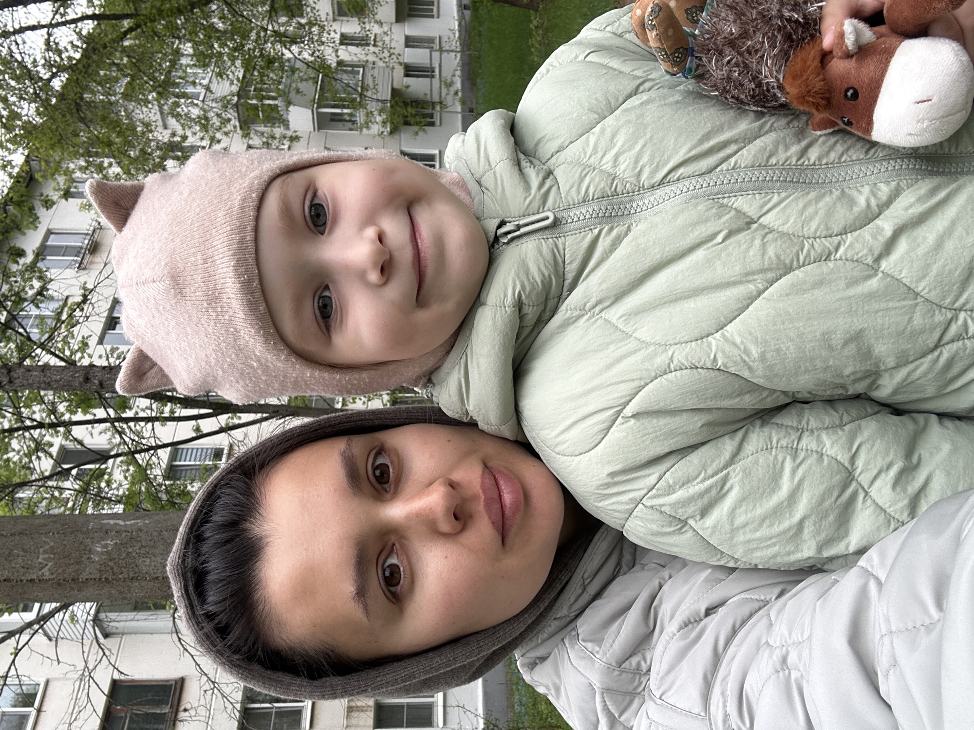 A woman in a grey hooded jacket is photographed holding a young child outdoors. The child, wearing a light pink hat with cat ears and a light green quilted jacket, holds stuffed animals. The background features apartment buildings and trees, suggesting a residential area in the city of Chaikovsky, Russia.