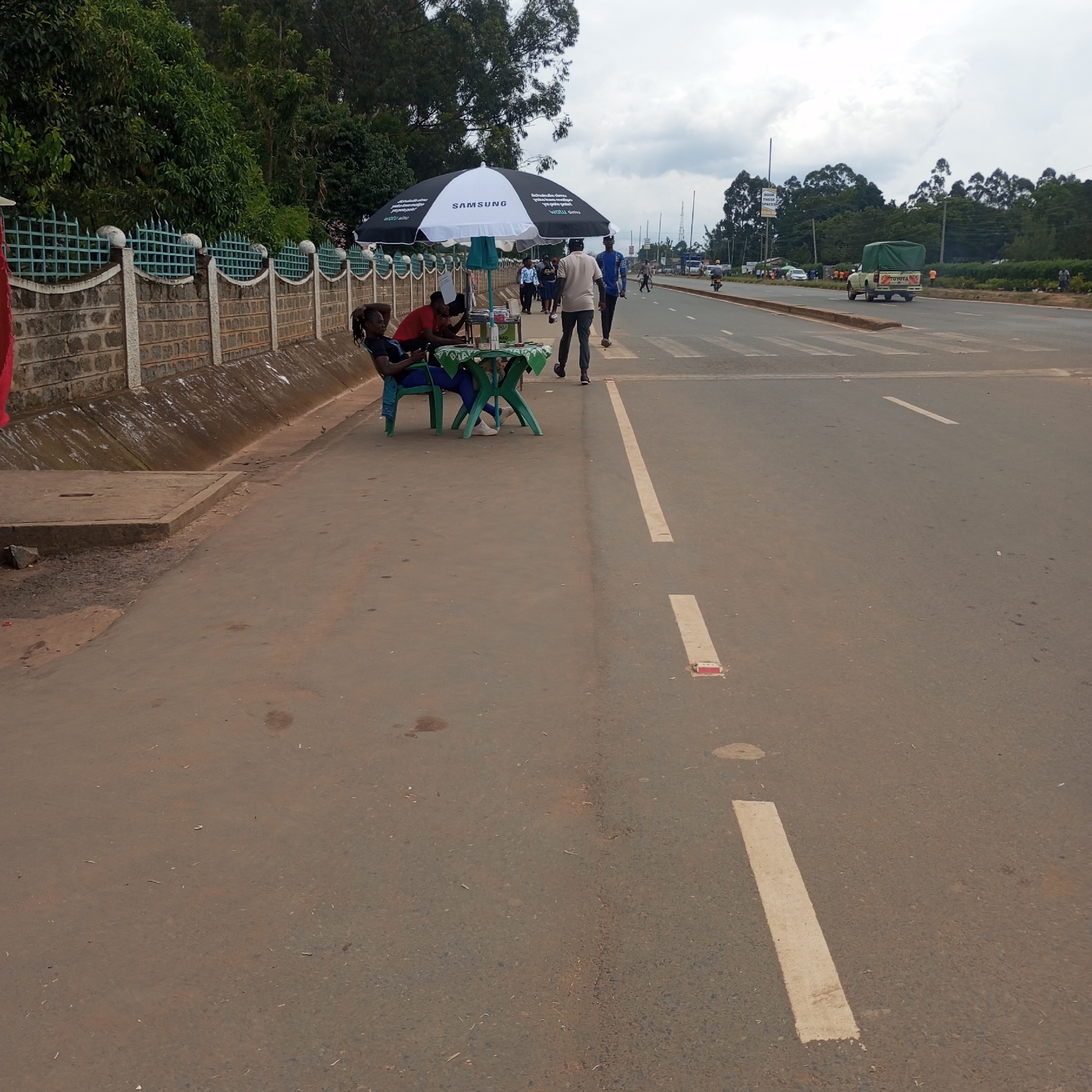 The image shows a roadside scene in Sheywe ward, Kenya.
A woman is seated on a green plastic chair at a green plastic table under a Samsung-branded umbrella. Other individuals are nearby.
A road runs parallel to the sidewalk with a white dashed line separating the lanes.
A white Toyota pickup truck is visible on the road. There are trees and a brick wall with a green metal fence in the background.