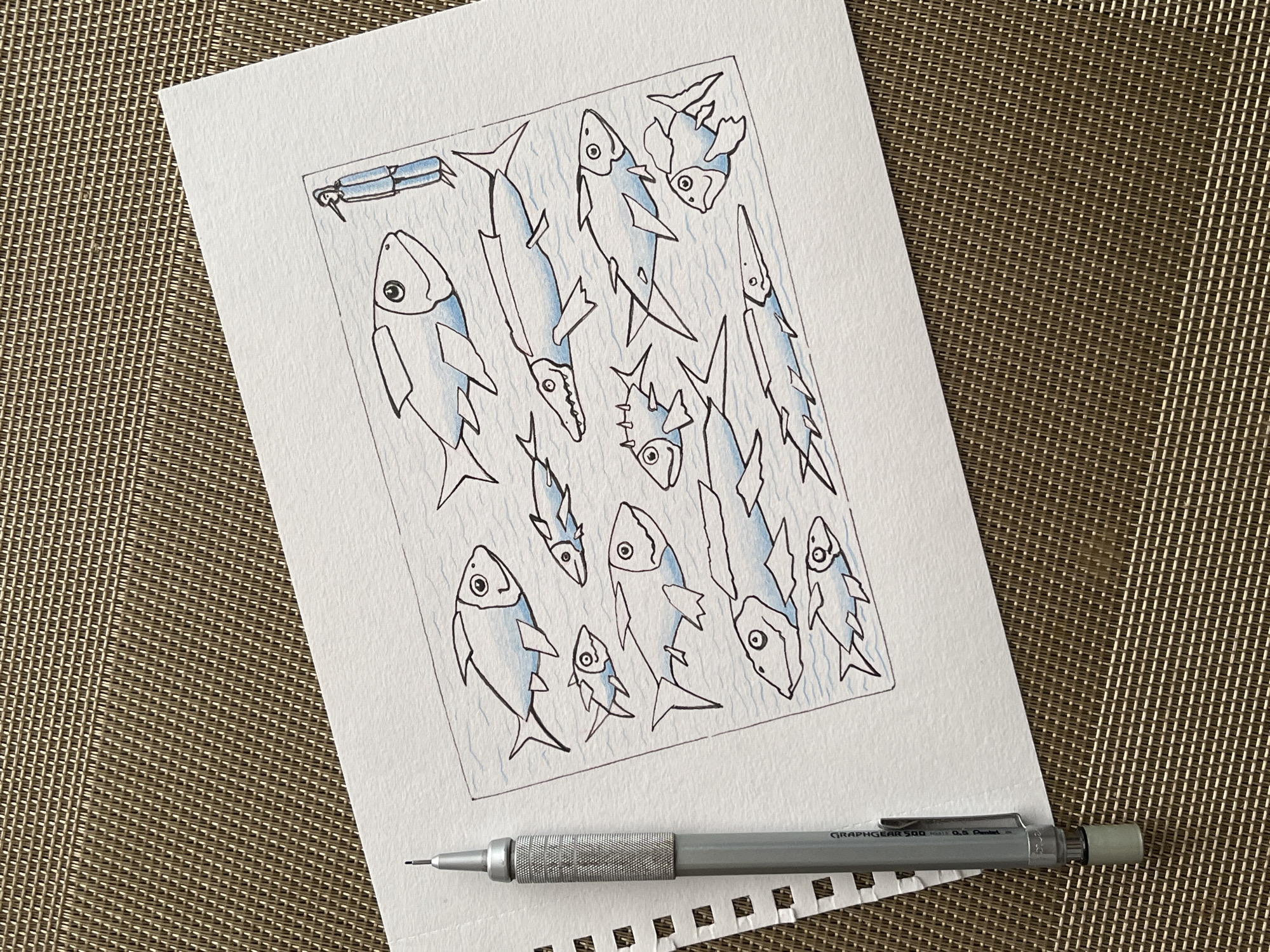 The image displays a drawing on white paper featuring a school of stylized fish and a small, dark-haired figure in a blue suit. The drawing is enclosed within a thin black border. The fish are rendered in simple black outlines with washes of light blue watercolor. They vary in size and pose, some appearing to swim in a school. The figure, positioned to the right of the fish, is depicted in a side profile, facing the fish. The paper rests on a woven brown mat. A mechanical pencil with a textured grip and a silver tip is visible on the left side of the image, positioned to the left of the drawing. The background of the mat appears to be a neutral woven texture. There is no indication of Krasnoyarsk, Russia, or any specific location within a city.