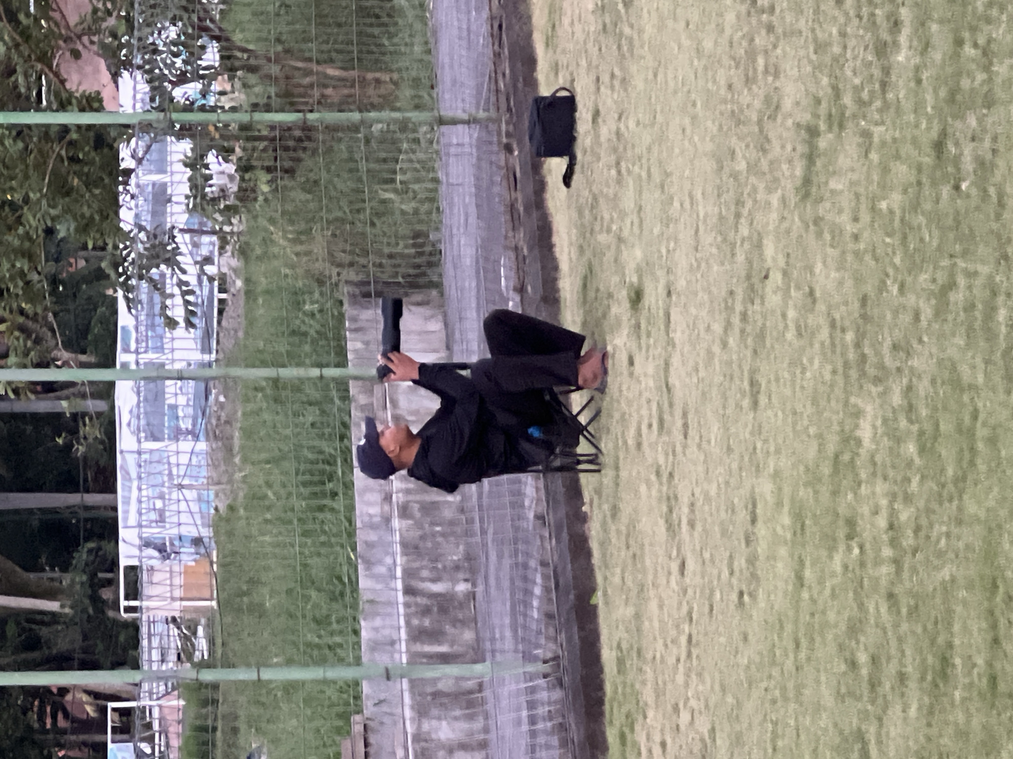 A man in black attire and a cap sits in a folding chair on a grassy area. He appears to be looking through binoculars or a similar device. A black bag sits beside him. A concrete wall and a green wire fence are behind him. In the background is a building or structure with scaffolding visible through the fence. The location is likely the Ubud District of Indonesia.