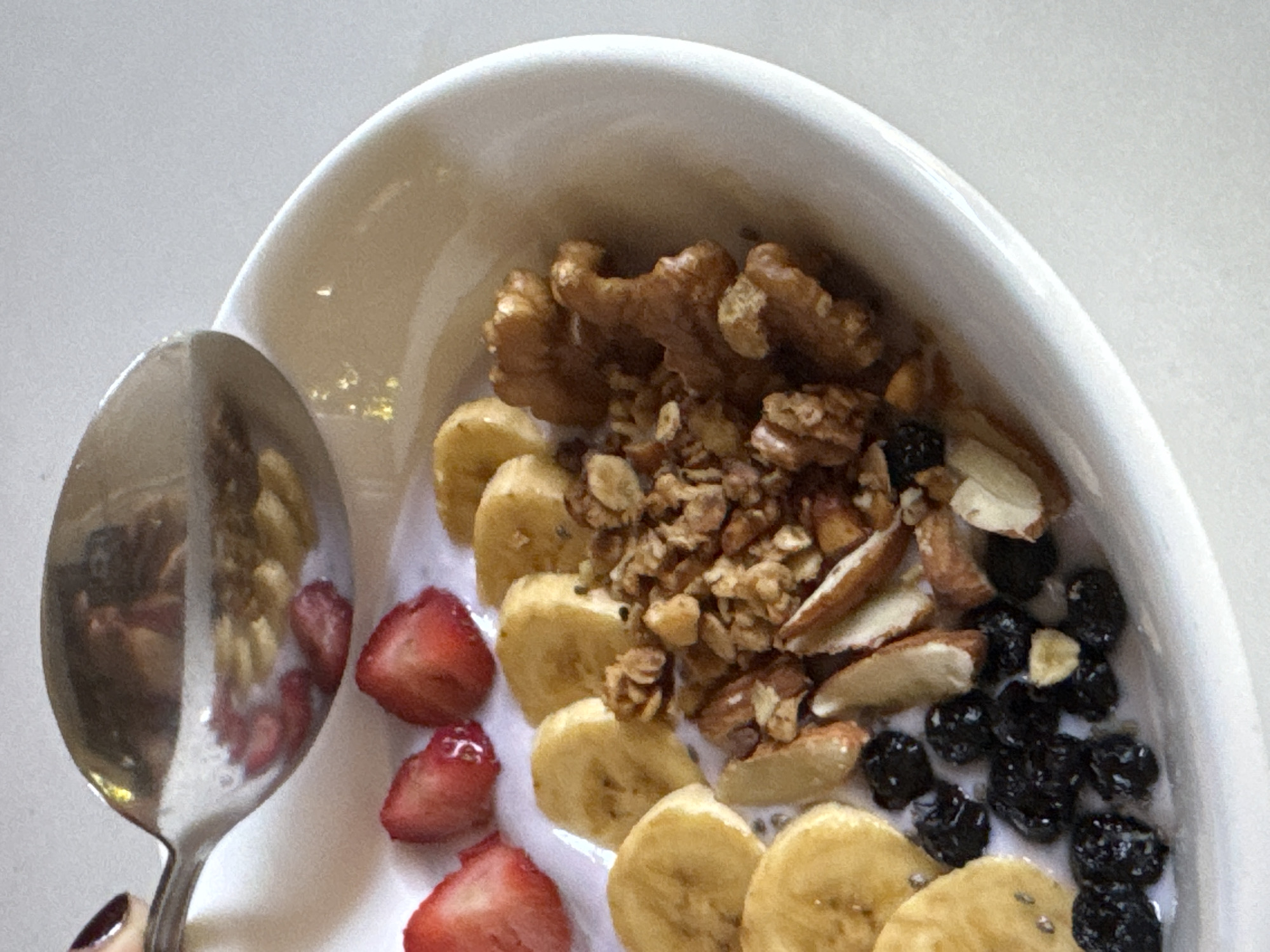 A close-up shot captures a white bowl filled with a healthy breakfast. Sliced bananas, strawberries, blueberries, granola, almonds, and walnuts are artfully arranged in the bowl, partially submerged in what appears to be yogurt or milk. A metal spoon, held by a hand with dark nail polish, hovers over the bowl, with a few pieces of strawberry visible on its surface. The background is a plain, light-colored surface, suggesting an indoor setting. There is no information available to determine the location in Davao City, Philippines, or any specific details about the people involved.