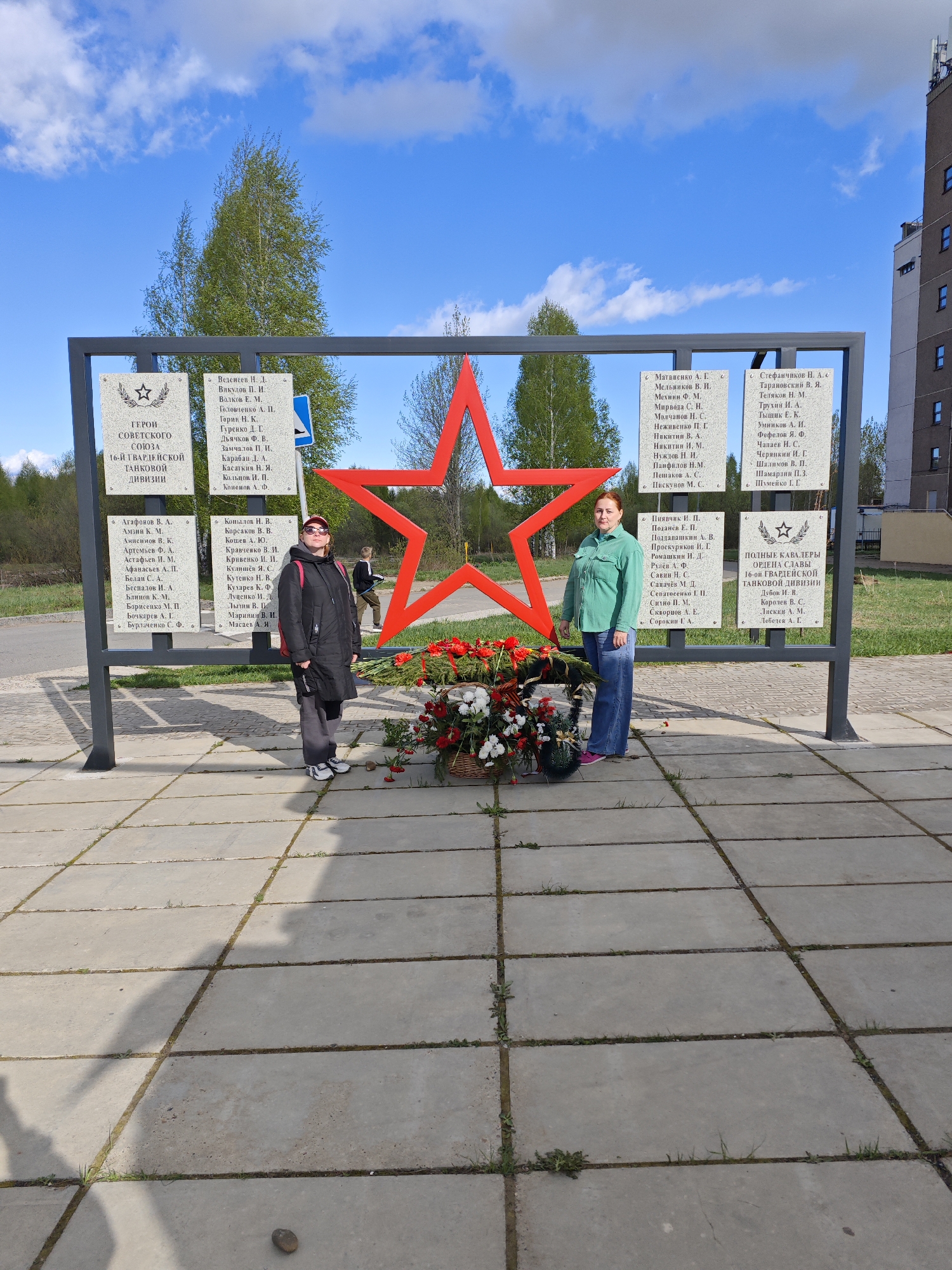 Here is a description of the image:
**Scene**: The image shows a monument in what is possibly a commemorative area, in the city of Марковский, Russia.
**Monument**: The monument consists of a gray metal frame holding several stone plaques with inscriptions on them, likely commemorating individuals and military units. A large red star is centered within the frame. A basket of red and white flowers sits at the base of the monument.
**People**: Two women are standing in front of the monument. The woman on the left is wearing a dark coat, gray pants, and sneakers. The woman on the right is wearing a green jacket, blue jeans, and athletic shoes.
**Environment**: The ground is paved with large concrete blocks. Trees are visible in the background, with a building partially visible on the right side. The sky is blue with scattered clouds.