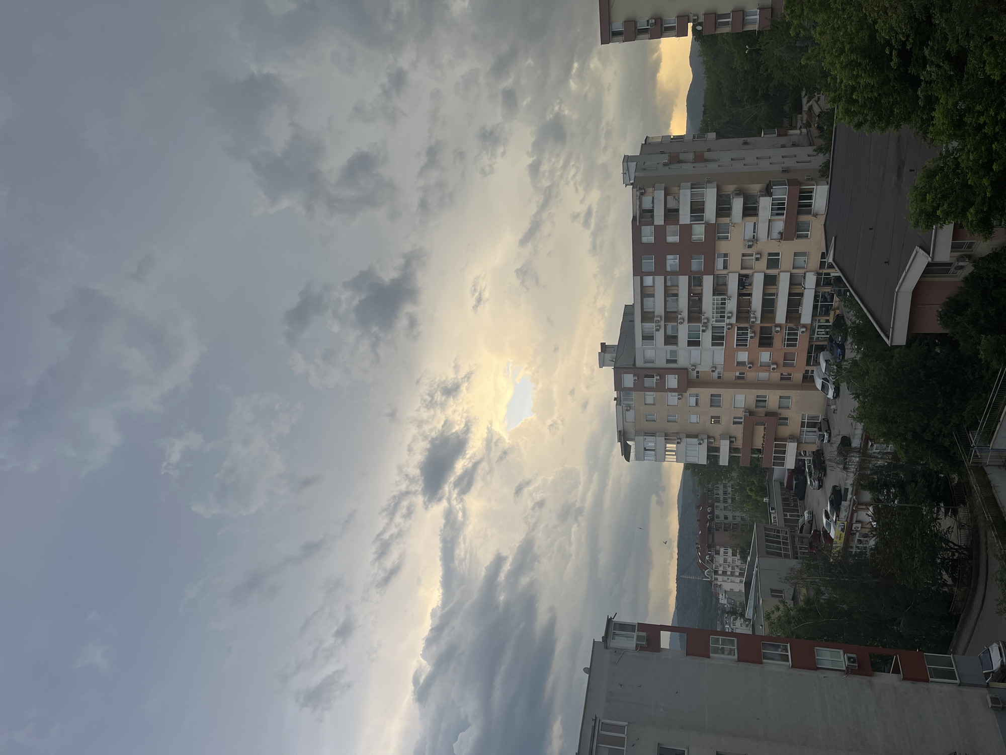 The image presents an urban scene under a partly cloudy sky, likely taken from a high vantage point. The central focus is a residential building with a mix of beige, brown, and white colors. The building has multiple floors and numerous windows, some equipped with air conditioning units and balconies. A variety of parked vehicles are seen in the adjacent space.
In the background, a mountain range is faintly visible, providing a natural backdrop to the urban environment. Part of the city Veliko Tarnovo, Bulgaria, has a generally gray tone, possibly due to the weather conditions.
There are trees with visible green foliage on the right edge of the frame. Additionally, there are other buildings in the immediate foreground and background, indicating a densely populated area. The sky is characterized by a combination of dark, ominous clouds and patches of brighter sunlight, creating a dynamic contrast.