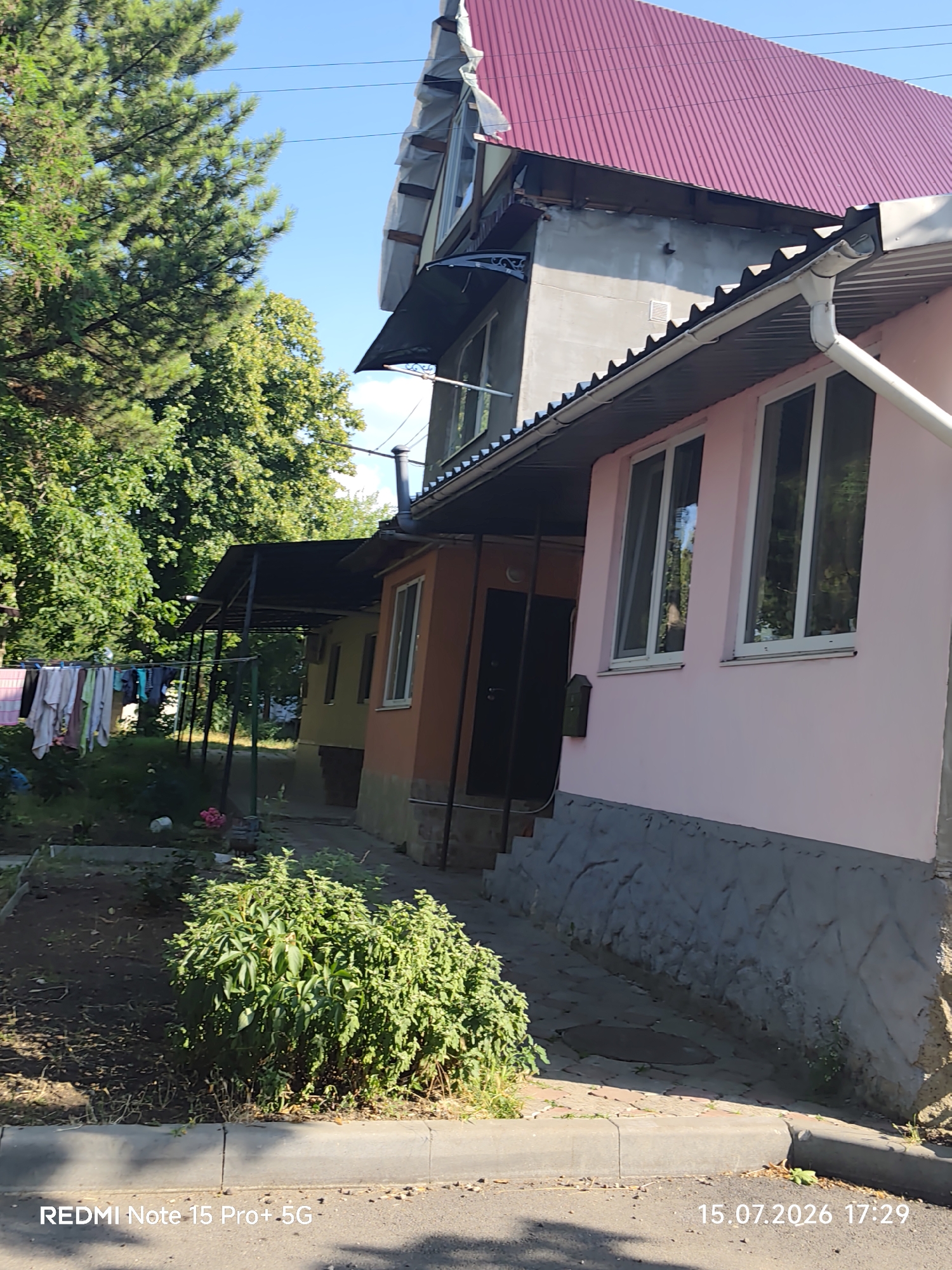 The image shows a residential building with a pink facade and a red corrugated metal roof, located in an outdoor setting. The building appears to be a two-story structure, with multiple windows visible on the front of the pink section. A covered walkway with a dark roof extends along the side of the building, supported by metal poles. Clothes are hanging on a clothesline in the background, suggesting residential activity. Lush green trees and foliage surround the building, indicating a natural or semi-urban environment. The ground in front of the building is paved with what appears to be cobblestones and concrete, with a raised curb visible along the edge of the street. The image also contains a timestamp in the bottom right corner, "15.07.2026 17:29".