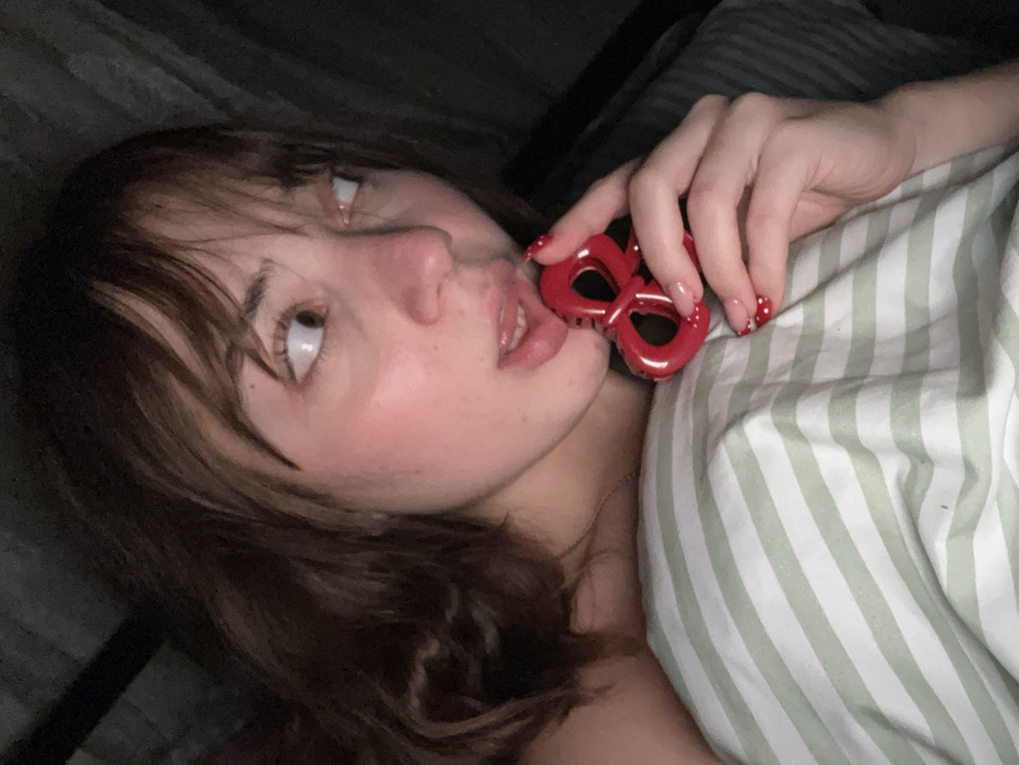 A young woman with brown hair and bangs, lying in bed under striped bedding, glances upward. She holds a red, figure-eight-shaped object near her mouth. Her fingernails are painted with a French manicure and red polka dots. The scene is dimly lit, creating a moody atmosphere.
