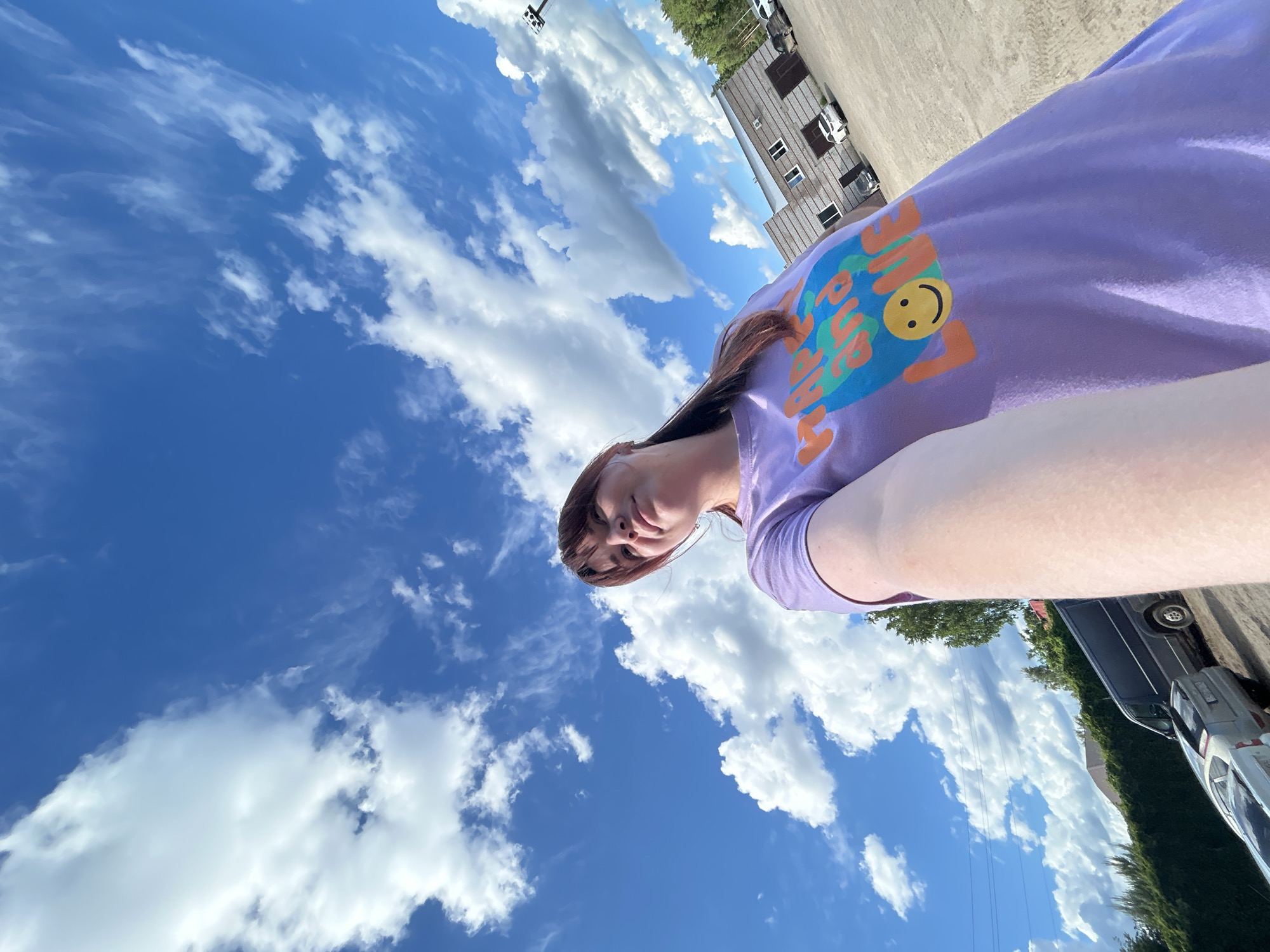 A low-angle, rotated perspective image features a person with shoulder-length brown hair and bangs, wearing a purple t-shirt with a colorful smiley face graphic. The person's head and torso are visible in the foreground.
The background is dominated by a vibrant blue sky with scattered cumulus clouds. In the upper right corner of the frame, a multi-story building with visible windows and a light-colored exterior is partially visible. At the bottom right, vehicles and dense green foliage are visible, suggesting a street or parking area.
Based on the visible building style and street context, it is plausible that the location is Chaikovsky, Russia, but it would require additional information to confirm.