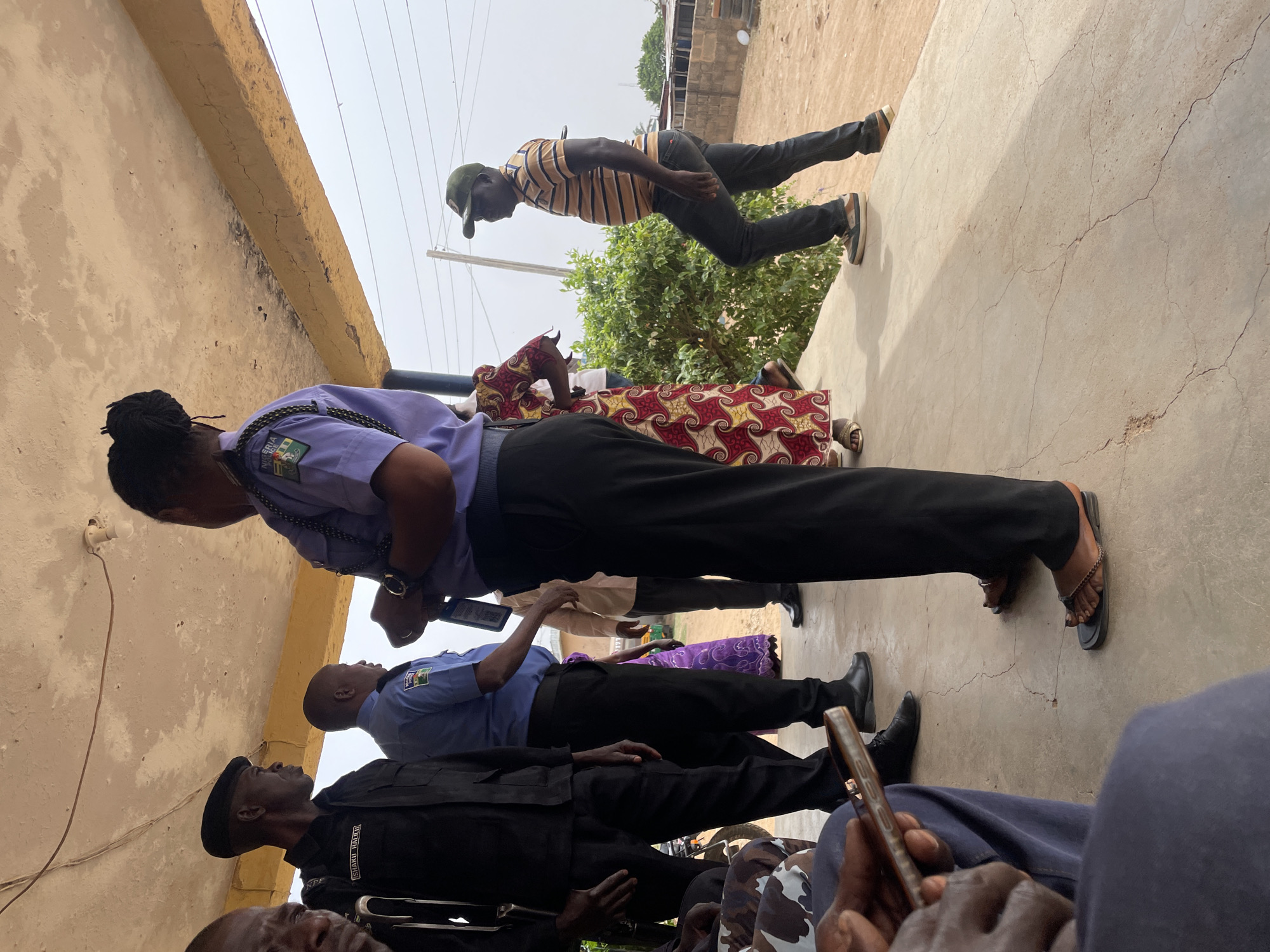 An outdoor scene in Jos, Nigeria, features several individuals, including uniformed police officers and civilians, gathered under a building overhang during bright daylight.
In the foreground, a female police officer stands prominently, dressed in a light purple uniform shirt with a "NIGERIA POLICE" crest visible on her left sleeve, black trousers, and black sandals. Her dark hair is styled in dreadlocks. She is looking downwards, possibly at an object in her hands. To her right, a male officer in a light blue uniform shirt with a similar "NIGERIA POLICE" crest, black trousers, and black shoes stands. Another male figure, closer to the foreground and dressed in a black uniform, black beret, and black trousers, has "SHAKA HAKURI" and "NPF" emblazoned on his uniform, and is looking upwards.
Further back, a civilian man in a yellow and black striped shirt, dark trousers, and a green cap is seen standing near green foliage. Partially visible behind the female officer is a woman wearing a red and patterned dress. In the immediate foreground, part of a person's body holding a smartphone is visible.
The setting is an open area with a concrete ground, showing numerous cracks. An adjacent light-colored, textured wall also exhibits cracks and wear. An exposed light bulb fixture with wiring is attached to the wall. Overhead wires are visible against a bright, overcast sky, contributing to the even lighting. Green bushes and glimpses of distant buildings suggest a residential or semi-urban environment. The individuals appear to be gathered, possibly engaged in conversation or waiting.