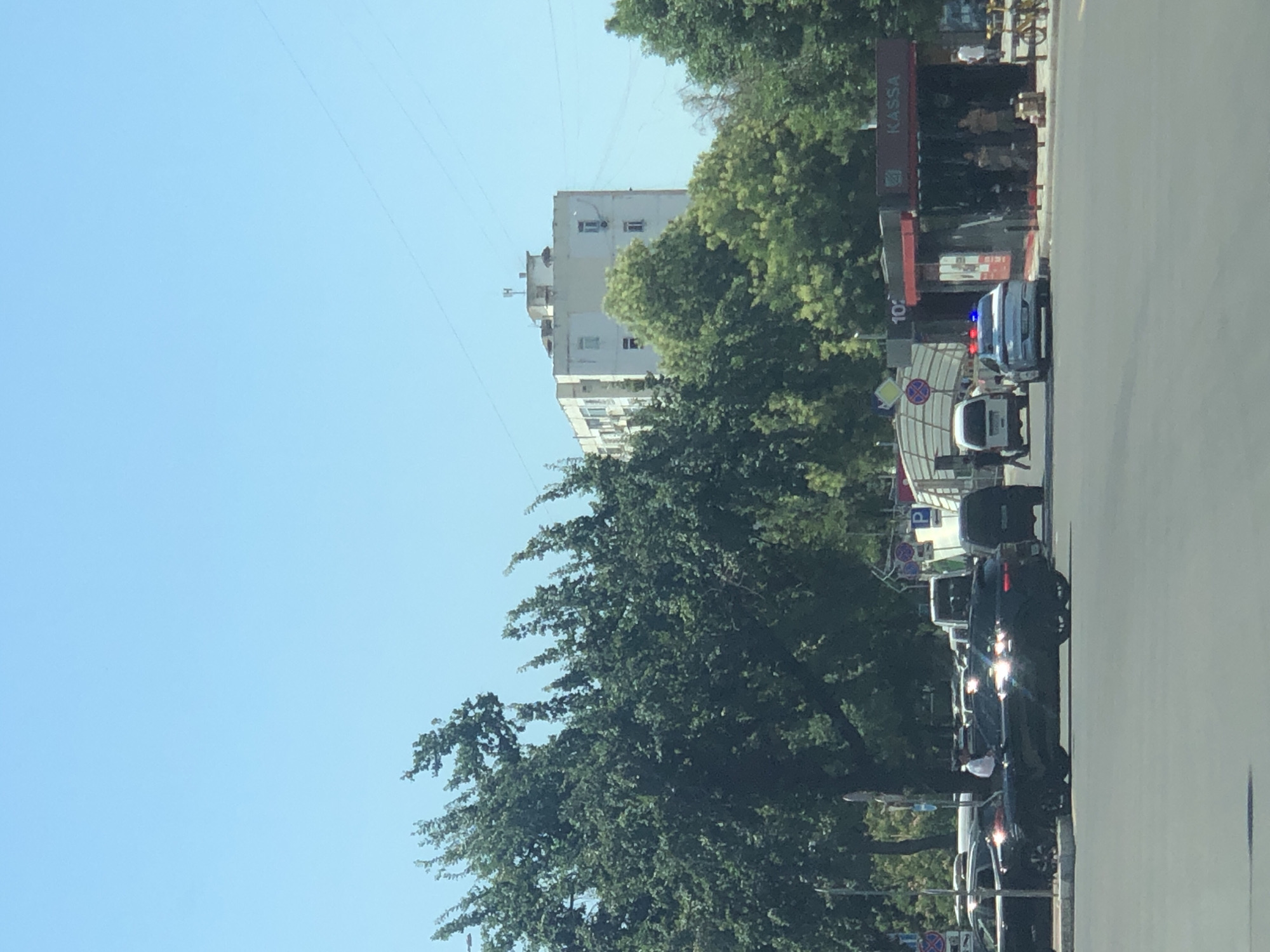 The image depicts a street scene in Tashkent, Uzbekistan, under a clear blue sky. In the foreground, a dark-colored SUV is parked, with a person in a white shirt standing next to it. Several other vehicles are visible, including a white minivan and a police car with flashing blue and red lights. Behind the vehicles, a building with multiple windows and an antenna on its roof stands out against the sky. Trees with lush green foliage line the street, creating a contrast with the urban structures. A red structure with the word "KASSA" visible suggests a ticket booth or similar establishment. The scene appears to be a typical day in the city, with traffic and pedestrian activity.