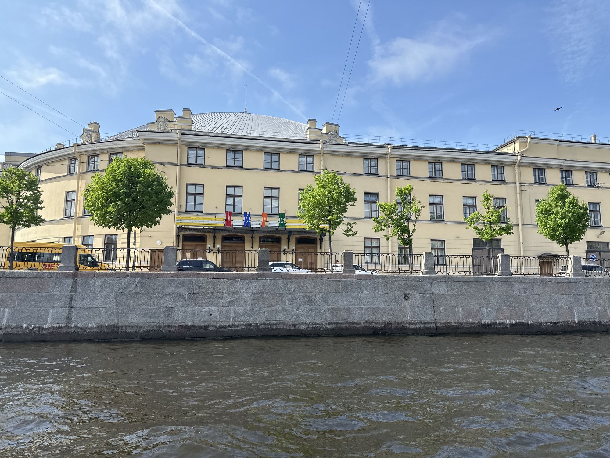The image depicts the Bolshoi Saint Petersburg State Circus building, located on the Fontanka Embankment in Saint Petersburg, Russia. The light yellow, multi-story building features a rounded facade and a dome-shaped roof. Trees line the embankment in front of the building, interspersed with a black wrought-iron fence atop a grey stone wall. Several vehicles are parked along the street. The Fontanka River fills the foreground, with visible water ripples. The sky is light blue with scattered clouds.