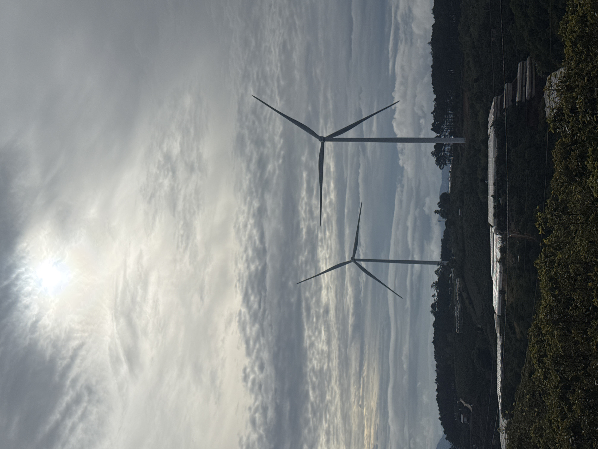Two wind turbines are situated in Phát Chi, Vietnam, beneath a sky filled with layered, overcast clouds. The sun shines dimly through the clouds, creating a diffused glow. The turbines are positioned near a lush, green hillside interspersed with what appears to be agricultural greenhouses. The terrain slopes gently upwards from the lower part of the frame.