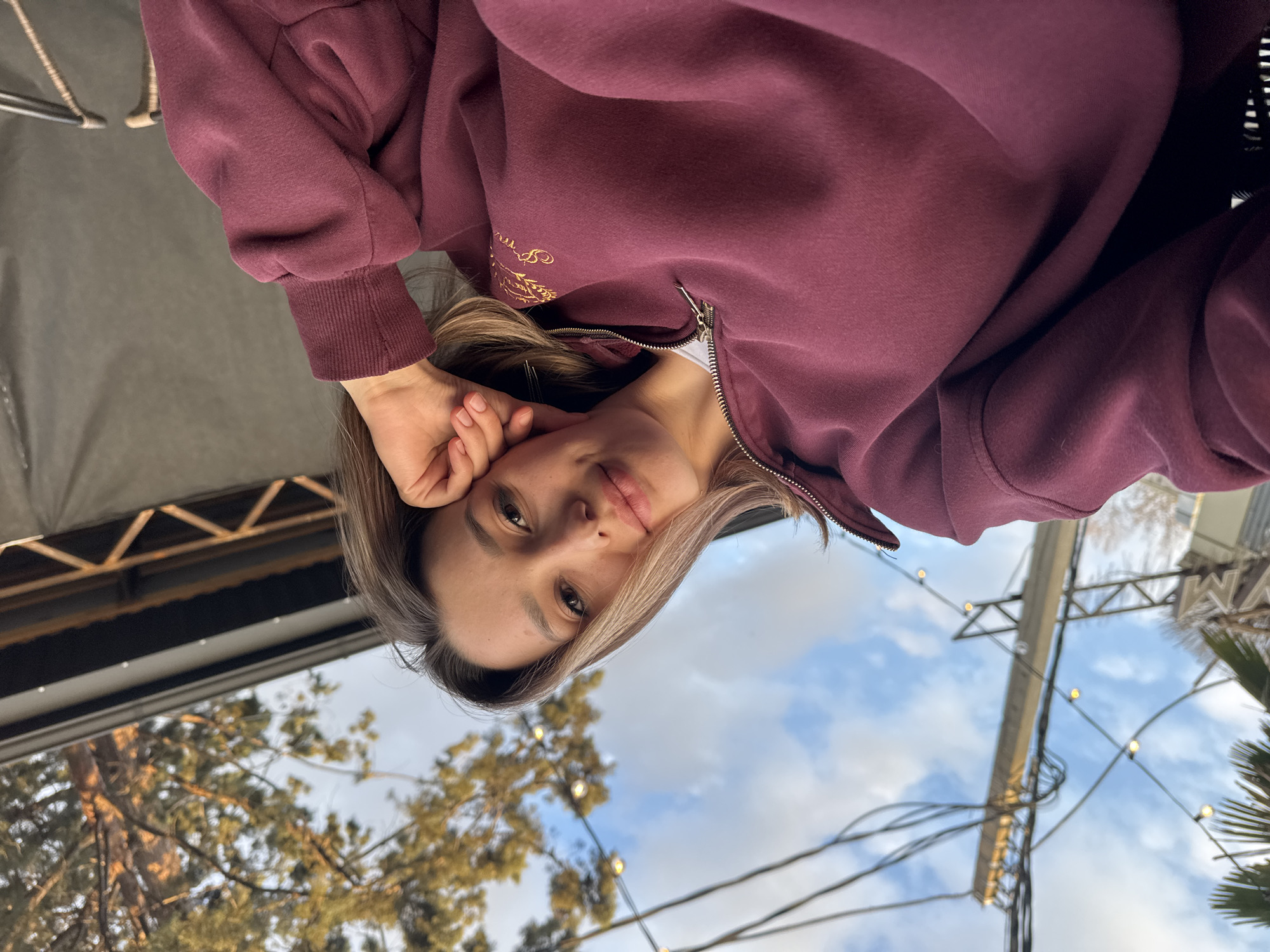 A casual, low-angle portrait features a young woman looking directly at the camera, with the image rotated 90 degrees counter-clockwise. She has light skin, dark eyes, and two-toned hair that transitions from darker roots to lighter, blonde ends. Her lips are slightly parted, and she has a calm, relaxed expression. Her left hand is gently resting near her face, revealing neatly manicured nails.
She is wearing a dark red or maroon zippered hoodie. A detail visible on the front of her hoodie is an intricate golden embroidered design, possibly text or a pattern, which is too blurry to be legible.
The background suggests an outdoor setting during daytime, possibly late afternoon, with a mix of natural and man-made elements. Behind her, a partially cloudy blue sky is visible, along with the tops of green and brown trees, likely conifers. Strewn across the sky and between structures are strings of small, glowing lights and various wires. Metal beams and structural elements, possibly from a canopy or building, are also present. The overall lighting is soft and natural.