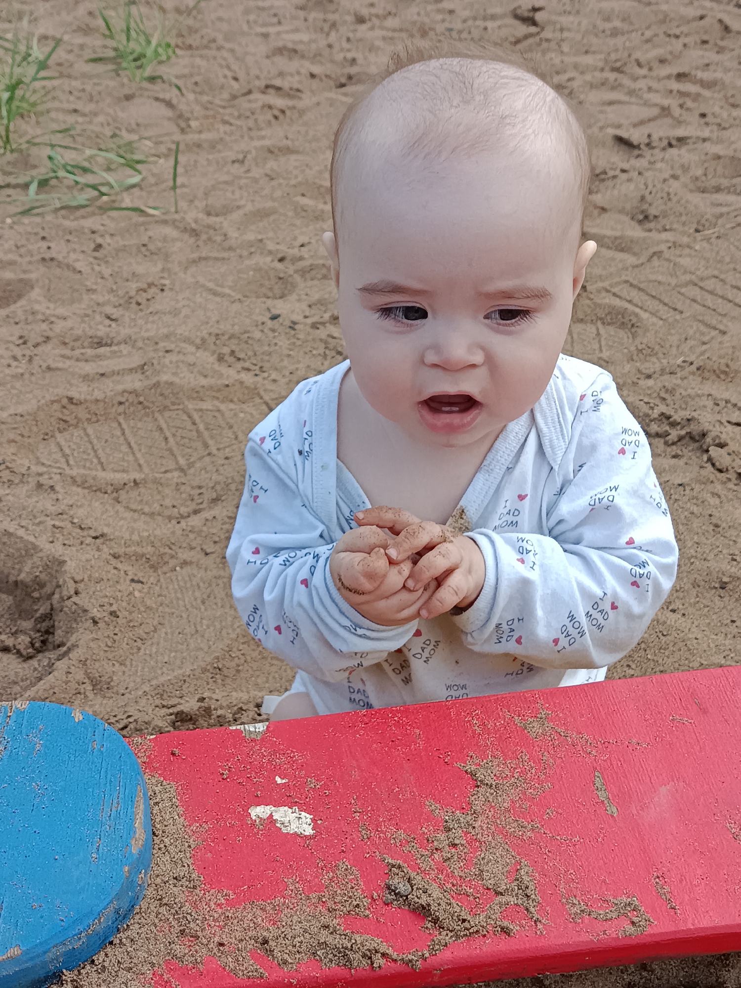 A baby sits in a sandbox. The baby is fair-skinned and bald with dark eyes and is wearing a white long-sleeved onesie with a pattern of the words "WOW MOM I LOVE DAD". The baby is holding clumps of sand in its hands. A blue wooden object and a red wooden object are in the foreground, covered in sand. The background consists of sand, with a few blades of grass visible. The location is the city of Chaikovsky, Russia.