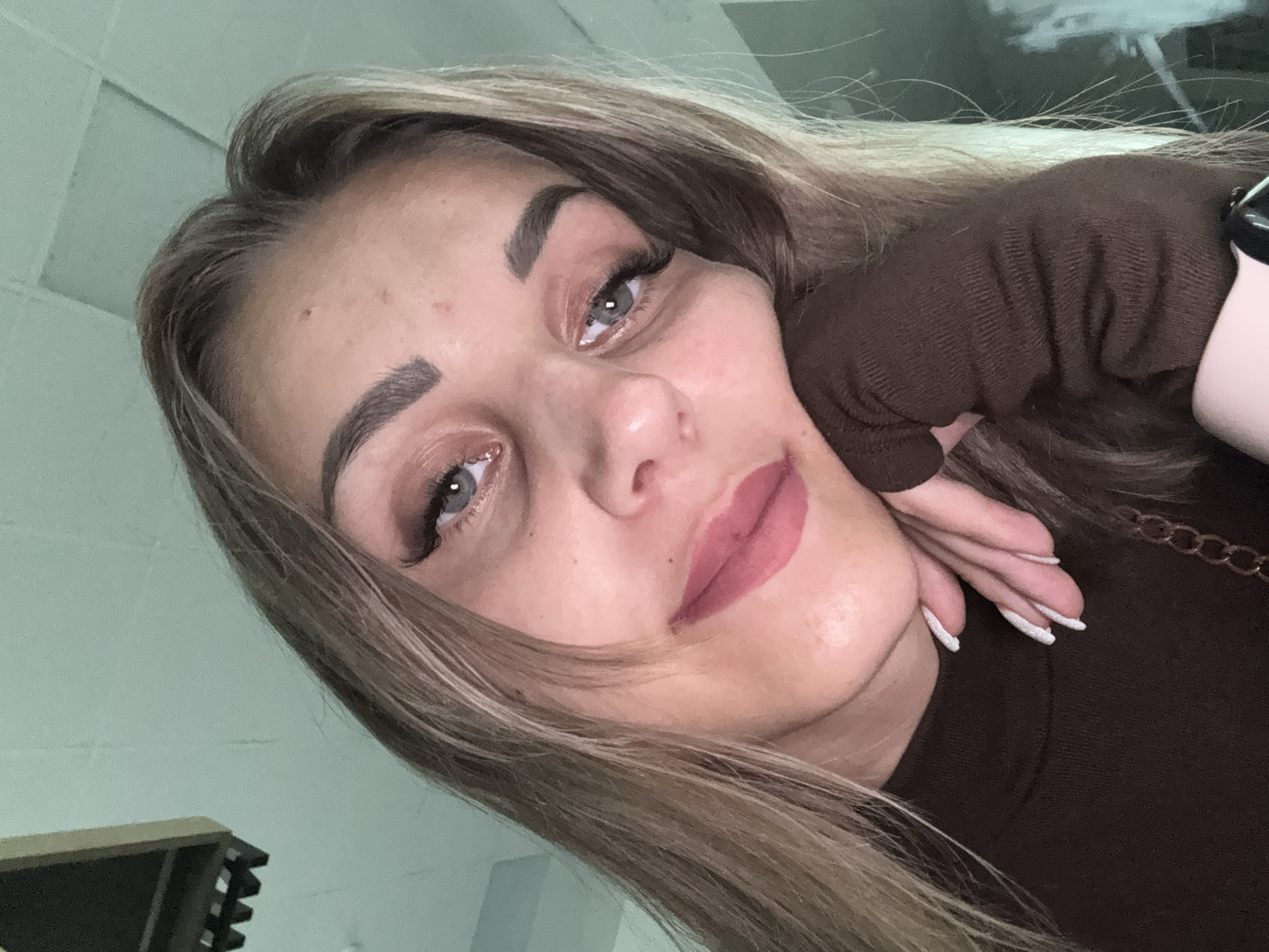 A close-up selfie shows a young woman with blonde hair and blue eyes resting her head on her hand. She is wearing a brown turtleneck sweater and has on makeup, including mascara, eyeshadow, and lipstick. Her nails are painted white and glittery. The background appears to be an indoor setting with a light green wall and a suspended ceiling fixture. No specific location in Yuzhno-Sakhalinsk, Russia, can be determined from the image.