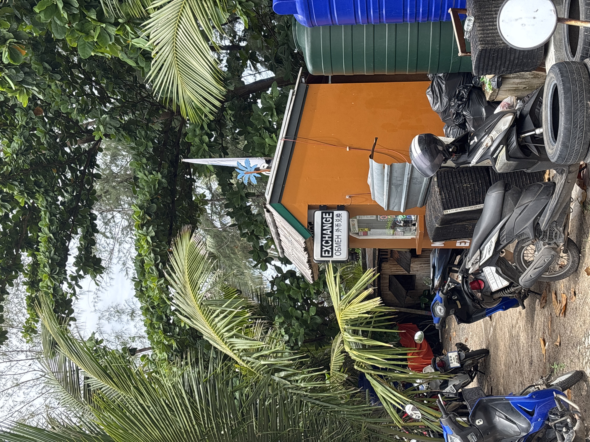 The image shows an exterior scene in Choeng Thale, Thailand. An orange building with a sign reading "EXCHANGE" in multiple languages is partially visible behind lush green foliage and palm trees. Several motorcycles are parked along a concrete path in front of the building. Miscellaneous objects, including tires, plastic water containers, and black bags, are piled beside the building. The sky is overcast.
