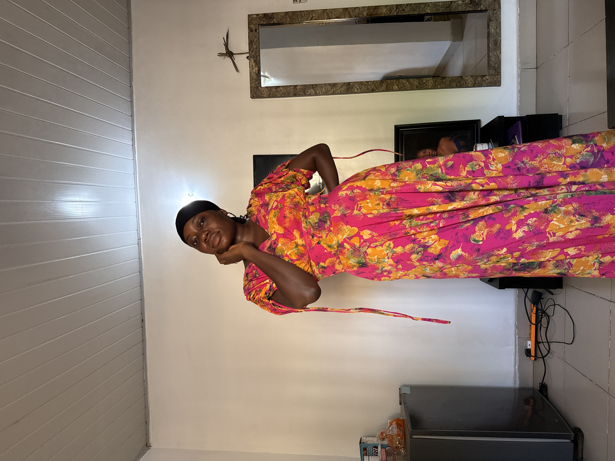 A young woman with dark skin is captured in a well-lit indoor setting, posing and looking directly at the camera with a soft, contemplative expression. She wears a vibrant, ankle-length maxi dress featuring a bold floral print in shades of fuchsia pink, bright yellow, orange, and green. Her hair is covered by a black headscarf, and she rests her right hand gently on her chin.
The background shows light-colored walls, with a section on the left featuring horizontal grey-and-white paneling. A large rectangular mirror with a mottled brown frame hangs prominently on the wall behind her. Above the mirror, a small, dark, artistic decoration resembling a bird or insect is affixed. To the right of the woman, a black-framed picture or photograph is visible, both directly behind her and reflected in the mirror. Below this, a dark grey or black chest freezer or refrigerator unit is situated, with an orange power strip and a cluster of cables on the wall behind it. Several packaged items, one bearing the text "ERS," are placed on top of the appliance. The scene appears to be a domestic interior, possibly a living space or kitchen, with ample light suggesting it is daytime.