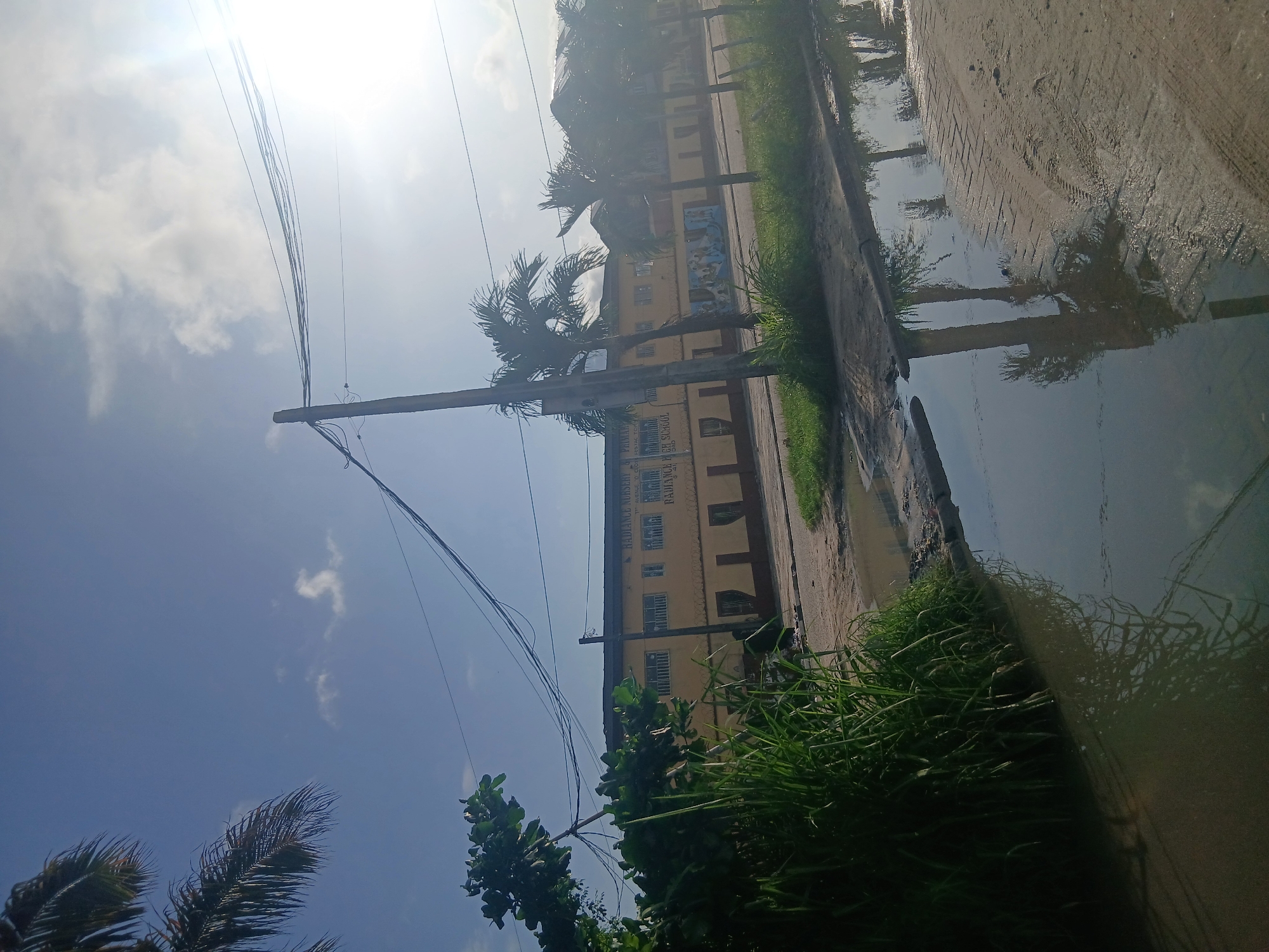 In Isunba, Nigeria, a street scene unfolds under a bright sky with scattered clouds. The sun glints brightly in the upper right corner. A multi-story building, painted a pale yellow, stands prominently. Text on the building reads "RADIANCE NURSERY & PRIMARY SCHOOL" and "RADIANCE HIGH SCHOOL". The building features numerous windows and a dark-colored roof. In front of the building, a line of palm trees can be seen. A utility pole with multiple wires stretches across the frame, extending from the bottom left to the top right. To the left of the building, dense green foliage and tall grass are visible. A muddy road with puddles of water occupies the foreground and right side of the image. Reflections of palm trees and the sky are visible in the water. The ground appears to be a mix of dirt and possibly some paving. Tire tracks are imprinted on the muddy surface.