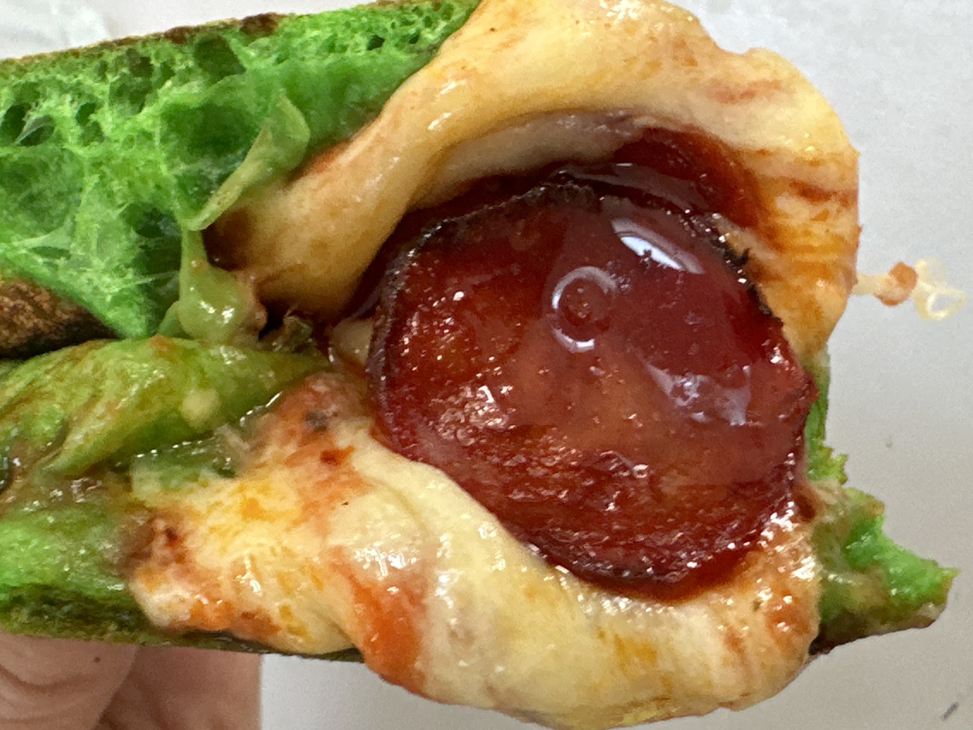 The image shows a close-up of a hot dog bun that has been dyed green, filled with melted cheese, sauce, and a slice of pepperoni. The bun appears to be slightly charred in places. The food is being held by a person's hand, though only a portion of the hand is visible. The background is a plain white surface. There is no discernible location in Moortown, Ireland, or any other specific environment.