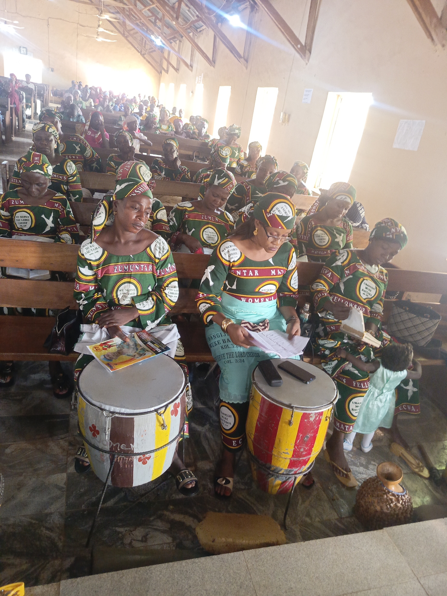 Captured indoors, likely within a church in Akwanga, Nigeria, the image shows a gathering of women seated in wooden pews. The majority are dressed in green, maroon, and white traditional attire with a distinctive circular emblem featuring the words "ZUMUNTAR MATA," "WE SERVE THE LORD CHRIST," and "WOMEN'S." Some women have matching headwraps. In the foreground, two women sit with painted drums bearing the markings “MEN” and “Me”. The drumheads have devices on them, likely smartphones, being used as sheet music for singing. A child stands close to one of the drummers. Other objects in view include a book, a handbag, an ornate vase, and discarded shoes. Natural light streams through the windows, illuminating the scene.