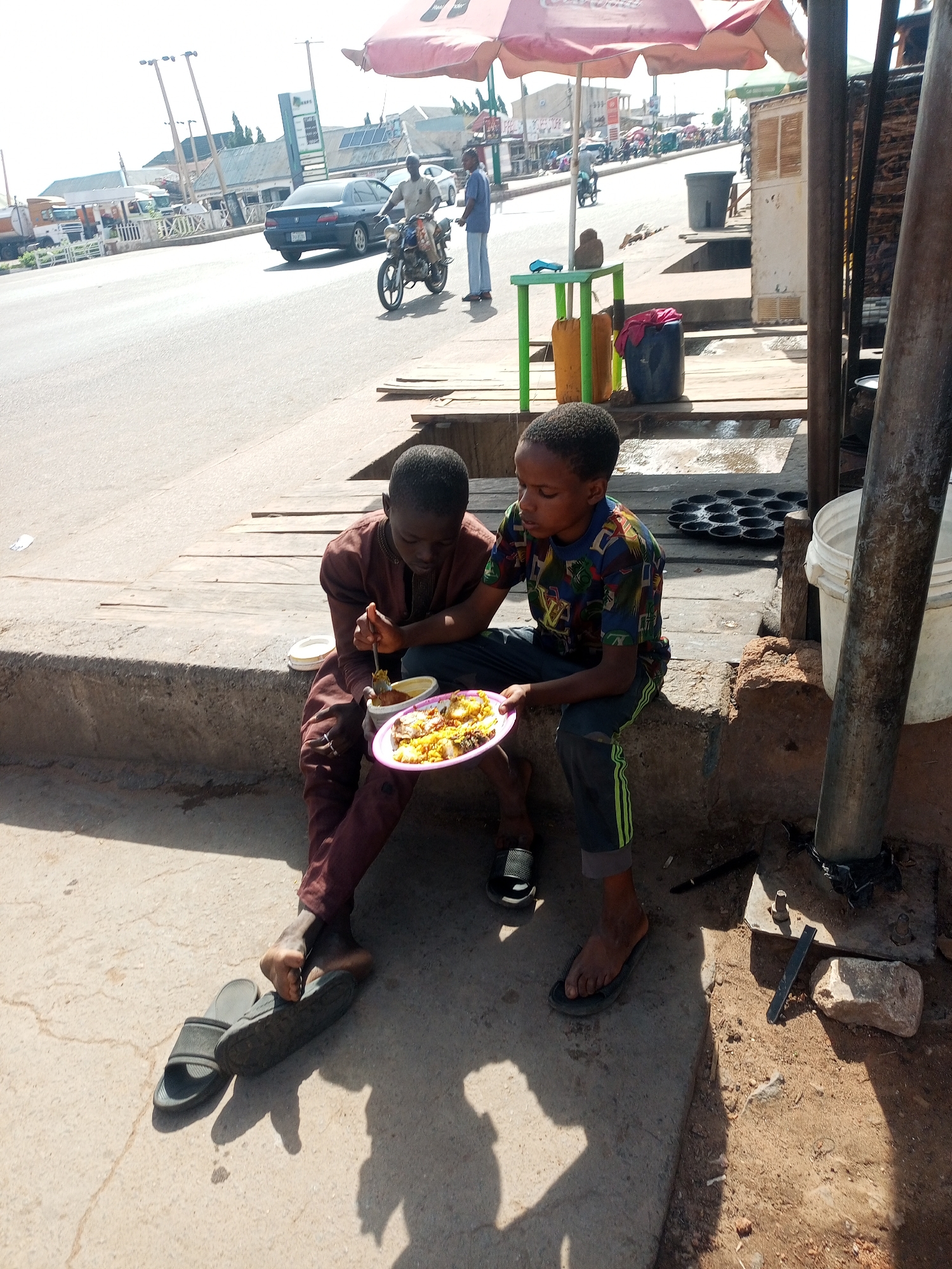 Two young boys are sharing a meal on a concrete ledge beside a busy street in Tumfure, Nigeria. The scene is illuminated by bright midday or early afternoon sunlight, casting distinct shadows. The boy on the left, dressed in a brown long-sleeved outfit, is eating from a small white bowl with a spoon, while his bare feet rest near a pair of black slides. The boy on the right, wearing a colorful patterned short-sleeved shirt and dark pants with green stripes, holds a pink plate filled with what appears to be rice and a stew or sauce. He is also barefoot, with one black slide visible near his foot.
The background shows a lively street scene with vehicular traffic, including a blue car and a motorcycle. Several adult figures are visible further down the road. The roadside features various elements such as a red-orange umbrella, a green plastic table with yellow containers, and other miscellaneous items suggesting a street-side vendor or market area. A black multi-cup baking tray and white plastic bucket are also visible near the boys. The setting appears to be an urban or semi-urban environment with buildings and power lines in the distance. The boys are engrossed in their meal, seemingly oblivious to the surrounding street activity. No clear text is legible in the image.