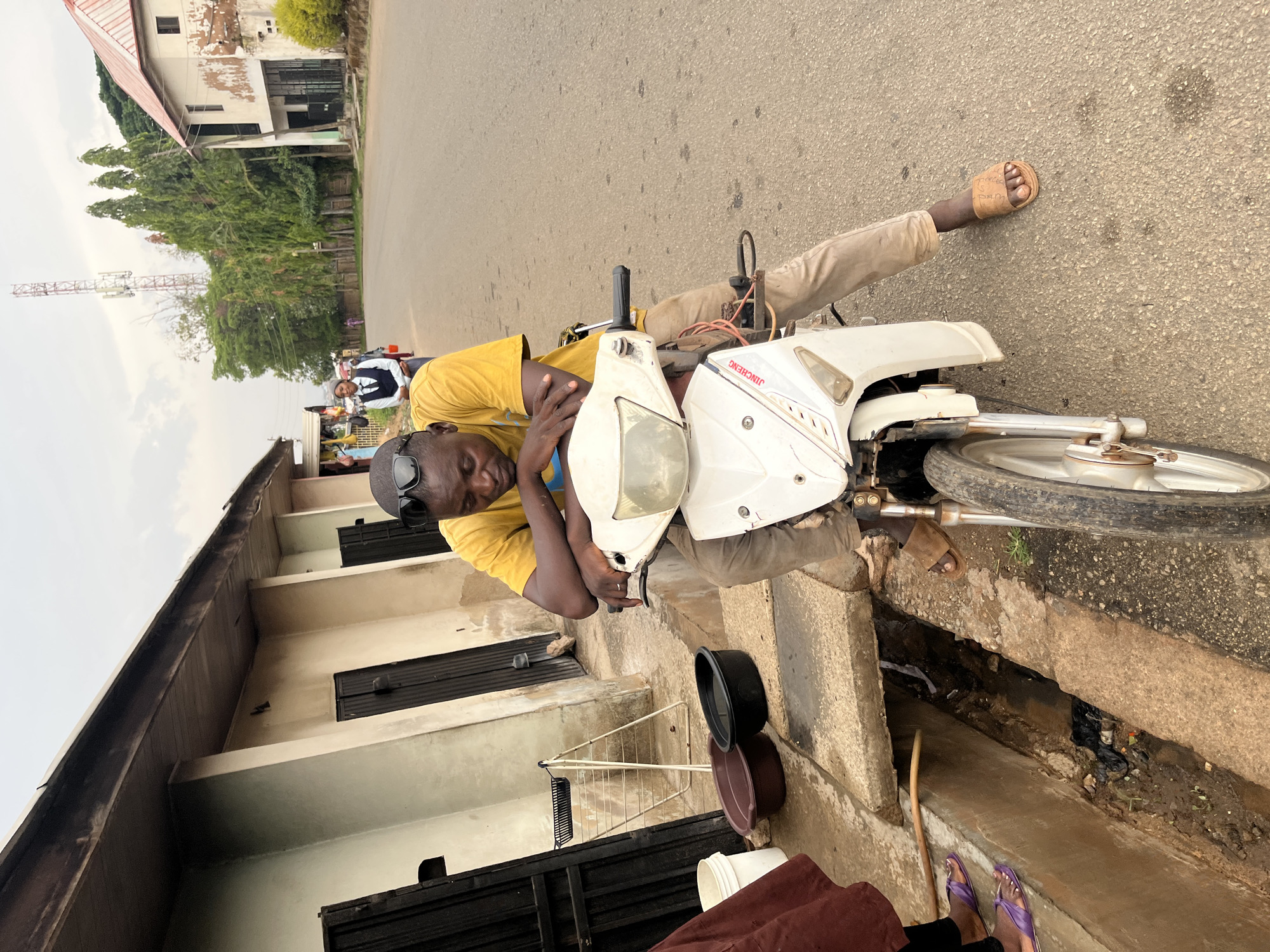 A man in a yellow shirt lies across a white motorcycle on a street. He wears sunglasses on his head and light-colored pants. The motorcycle is parked partially on the sidewalk next to a drainage ditch. A building is visible in the background on the left side of the image. A communications tower is visible further in the background to the left. Other people are also visible in the distance. The location is Unknown, Unknown.