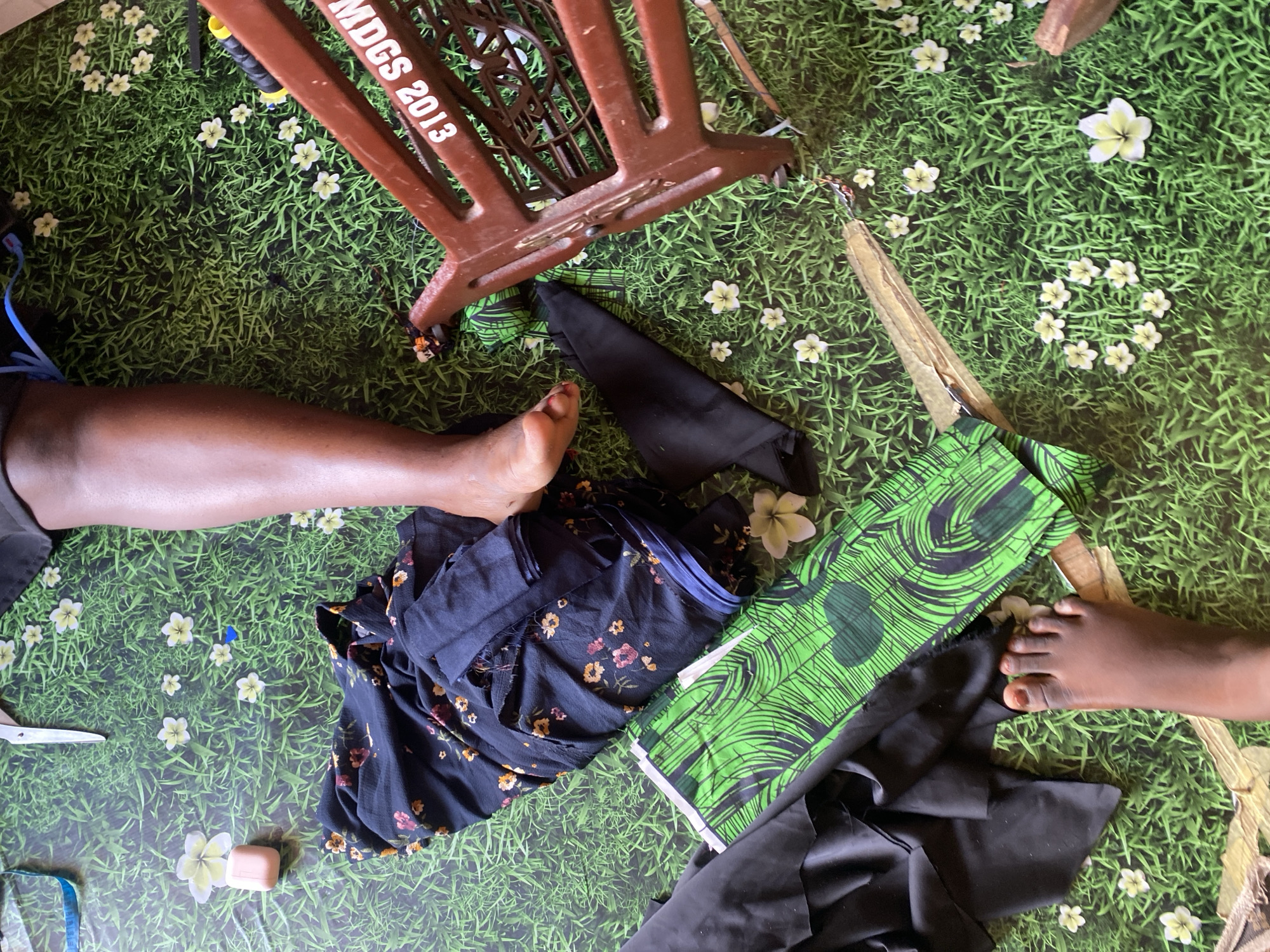 The image shows a top-down view of a person's legs and feet on a green, grass-patterned floor. To the left of the frame, there is a pile of dark blue fabric with a floral pattern and a piece of black fabric. A bright green fabric with a black leaf pattern is placed on top of the black fabric. In the upper right quadrant, a portion of a red sewing machine is visible, with the text "MDGS 2013" printed on it. A person's right leg is bent, with the foot resting near the fabric. Another foot is visible in the lower left portion of the image. The overall scene suggests a domestic environment where sewing is taking place. The location is identified as Jauro Yinu, Nigeria.