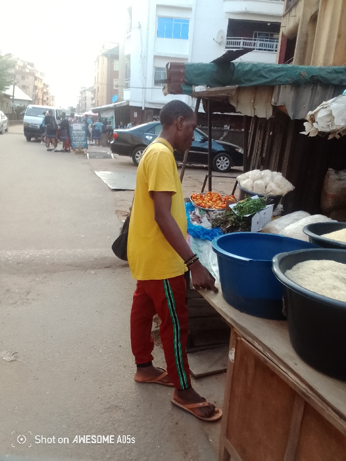 The image depicts an active daytime street market scene in Onitsha, Nigeria. In the immediate foreground, a dark-skinned male vendor stands in profile, facing left. He wears a yellow short-sleeved t-shirt, red sweatpants with vertical green and white stripes, and tan flip-flops. A dark bag is slung over his left shoulder, and multiple beaded bracelets are visible on his right wrist. His right hand is positioned on a wooden market stall.
The market stall, constructed from wood with a green tarp and corrugated metal roofing, displays various goods. These include a silver bowl containing small red spherical vegetables (consistent with tomatoes), bundles of green leafy vegetables, several white items packaged in clear plastic bags, a blue plastic basin, and a black plastic basin filled with a light-colored granular substance (consistent with rice).
Behind the vendor and stall, an asphalt road extends into the distance. A black sedan is visible parked or moving slowly on the road, with a white van and other vehicles further down the street. Multiple pedestrians are dispersed along the road and pavement. Multi-story urban buildings, some featuring blue-tinted windows and external satellite dishes, line both sides of the street.