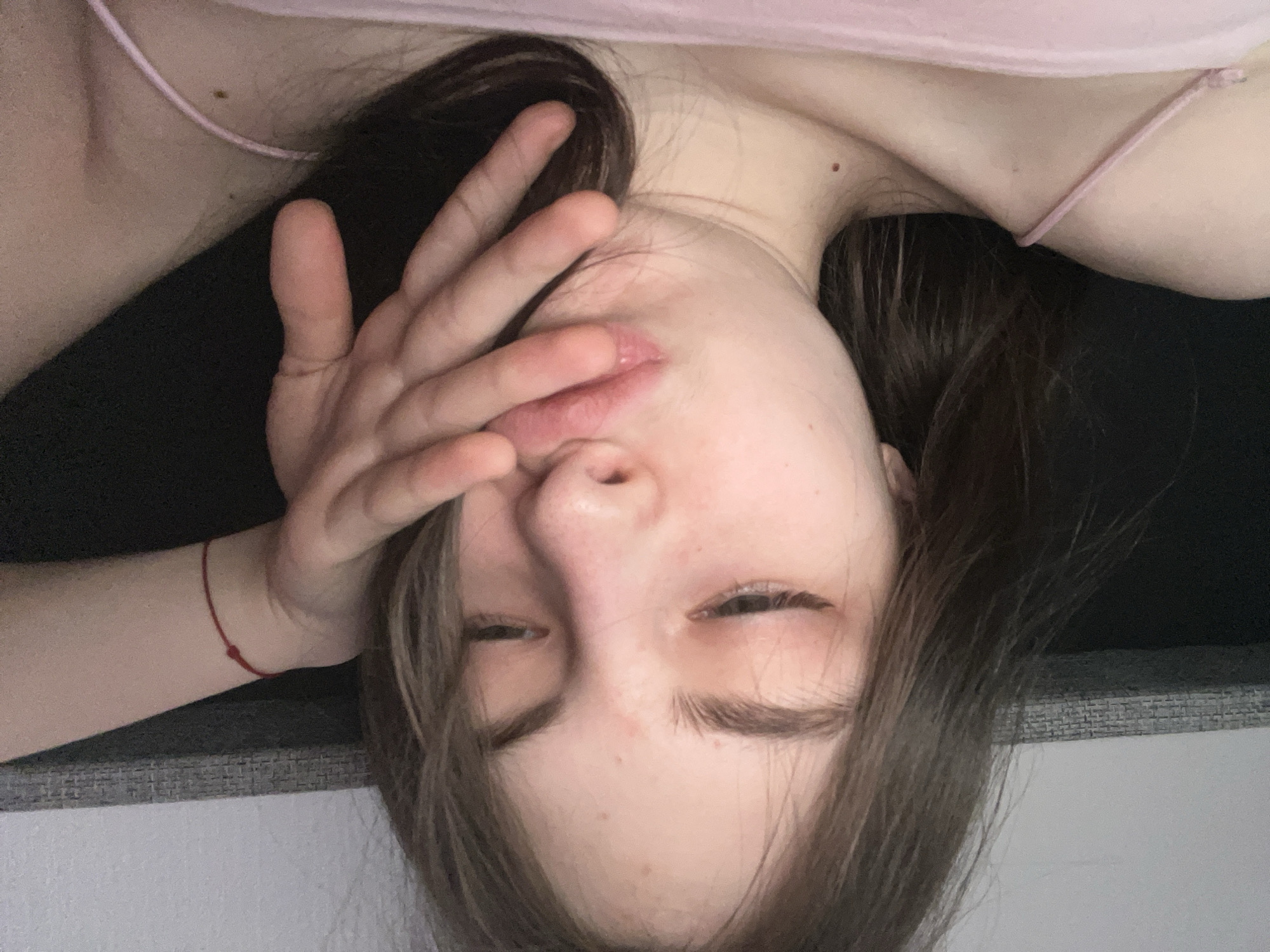 A woman lies on a grey and black surface, possibly a couch or bed, in an indoor setting. She wears a light pink top with thin straps. She rests her left hand on her face, partially covering her mouth and nose. The woman has dark brown hair. A red string bracelet is visible on her left wrist. It is impossible to determine her exact location and whether or not she is in Saint Petersburg, Russia based solely on the image.