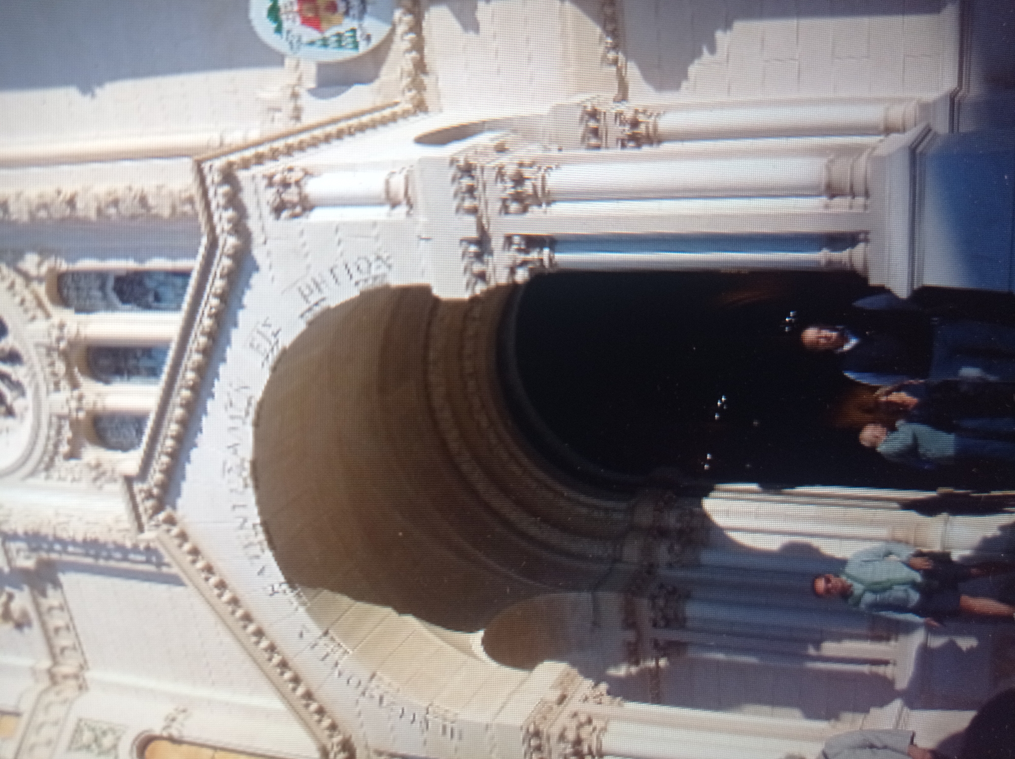 The image displays the facade of a large, light-colored building, likely a church or cathedral, with an arched entrance. The architecture features ornate carvings and pillars. In the foreground, several individuals are visible. One person, wearing sunglasses, a light blue shirt, and dark shorts, stands to the left. Another individual, wearing a dark vest over a blue shirt, stands to the right. In the background, indistinct figures can be seen within the arched entryway. The overall scene suggests an outdoor setting in front of a significant historical or religious structure. The location in the city of Mukachevo, Ukraine, is not discernible from the visual content of the image itself.