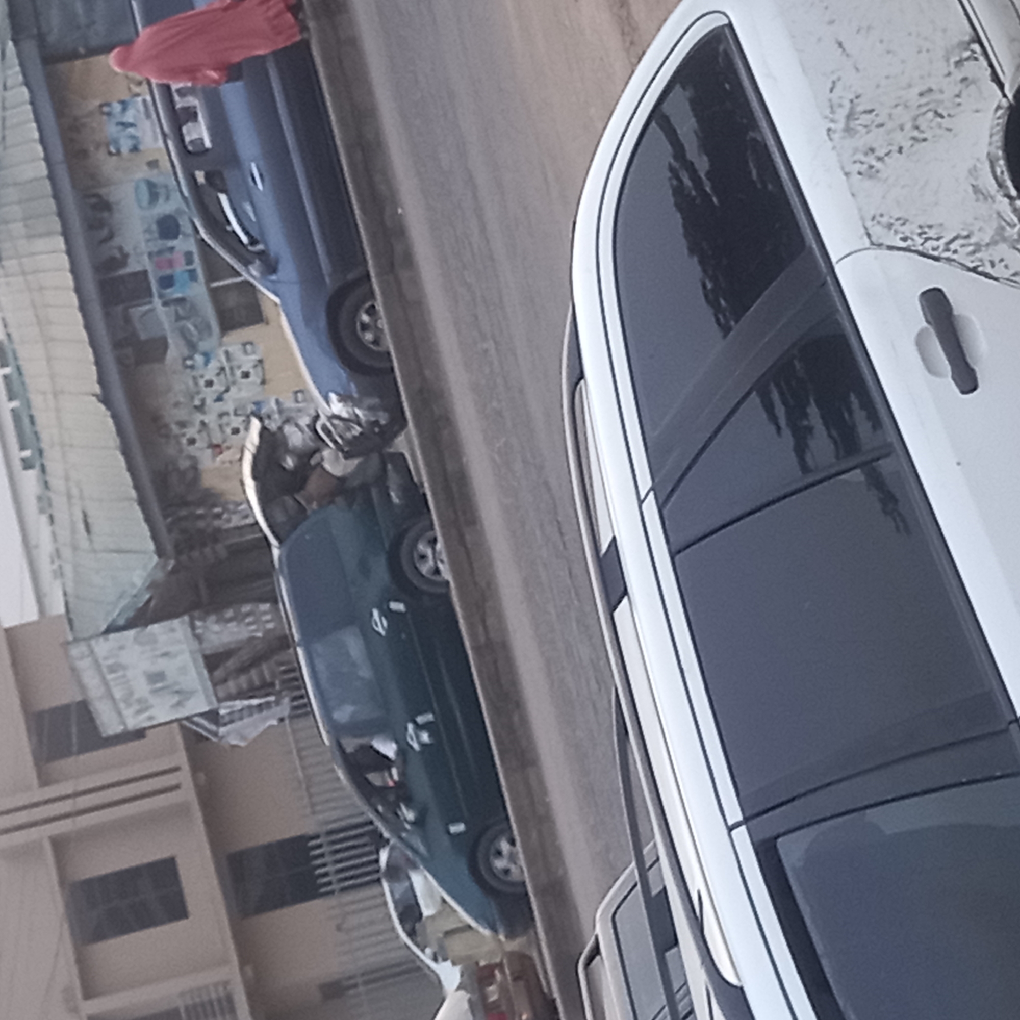 The image depicts a street in Dadi Riba, Nigeria, featuring several parked cars. A white vehicle is prominently visible on the right side of the frame, with a partially damaged roof. Adjacent to it, and further down the street, a dark green car is also parked. A blue car is also parked on the street. In the background, a building and a sign are visible, along with pedestrians on the sidewalk. The image is somewhat blurry, and the angle is tilted.