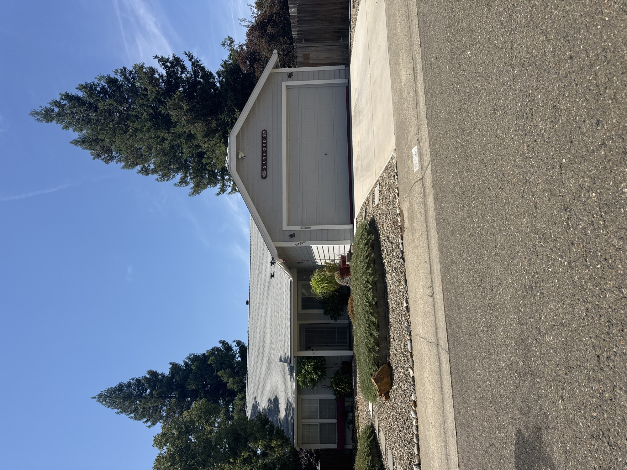 A grey, single-story house with a grey roof and a light grey garage door is visible from the street in Citrus Heights, California. The house has white trim around the windows and a dark red trim at the bottom of the garage door. A sign above the garage door reads "SANFORD" in black lettering. The house is surrounded by landscaping, including gravel, bushes, and a large rock in the front yard. Two tall evergreen trees are located on either side of the house, and a wooden fence is visible on the right side of the property. The sky is clear and blue. A paved driveway leads to the garage. The street in front of the house is asphalt.