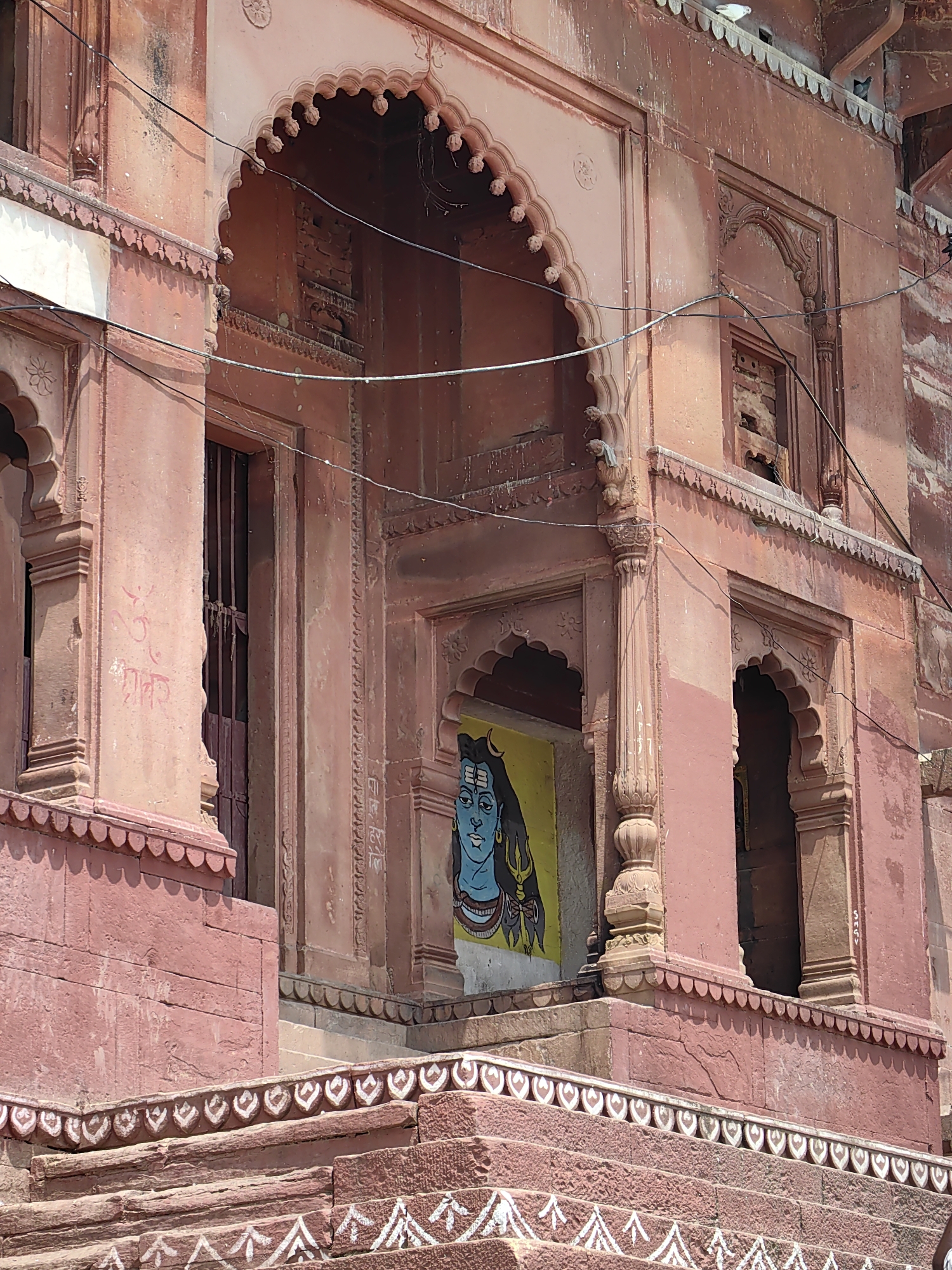 A medium shot captures the exterior of a building in Varanasi, India, with a focus on a richly decorated facade and a painted image. The architecture features ornate arches, intricate carvings, and a stepped base. The building appears to be constructed from reddish-brown stone, showing signs of age and weathering.
Dominating the center of the frame is a painting of Lord Shiva, depicted with blue skin, long dark hair, and a third eye on his forehead, rendered in a stylized, almost cartoonish manner against a bright yellow background. This painting is positioned within an arched alcove.
To the left of the Shiva painting, a dark-colored, barred doorway is visible. The wall to the left of this doorway has faint red script and an "Om" symbol painted on it. Above the doorway and arches, electrical wires are strung across the facade, partially obscuring the details of the upper levels of the building.
The foreground of the image shows a series of wide, stone steps leading up to the building. These steps are adorned with white decorative patterns, including arrow-like symbols and heart-shaped motifs. The overall impression is one of ancient architecture infused with vibrant religious art, typical of the spiritual landscape of Varanasi.