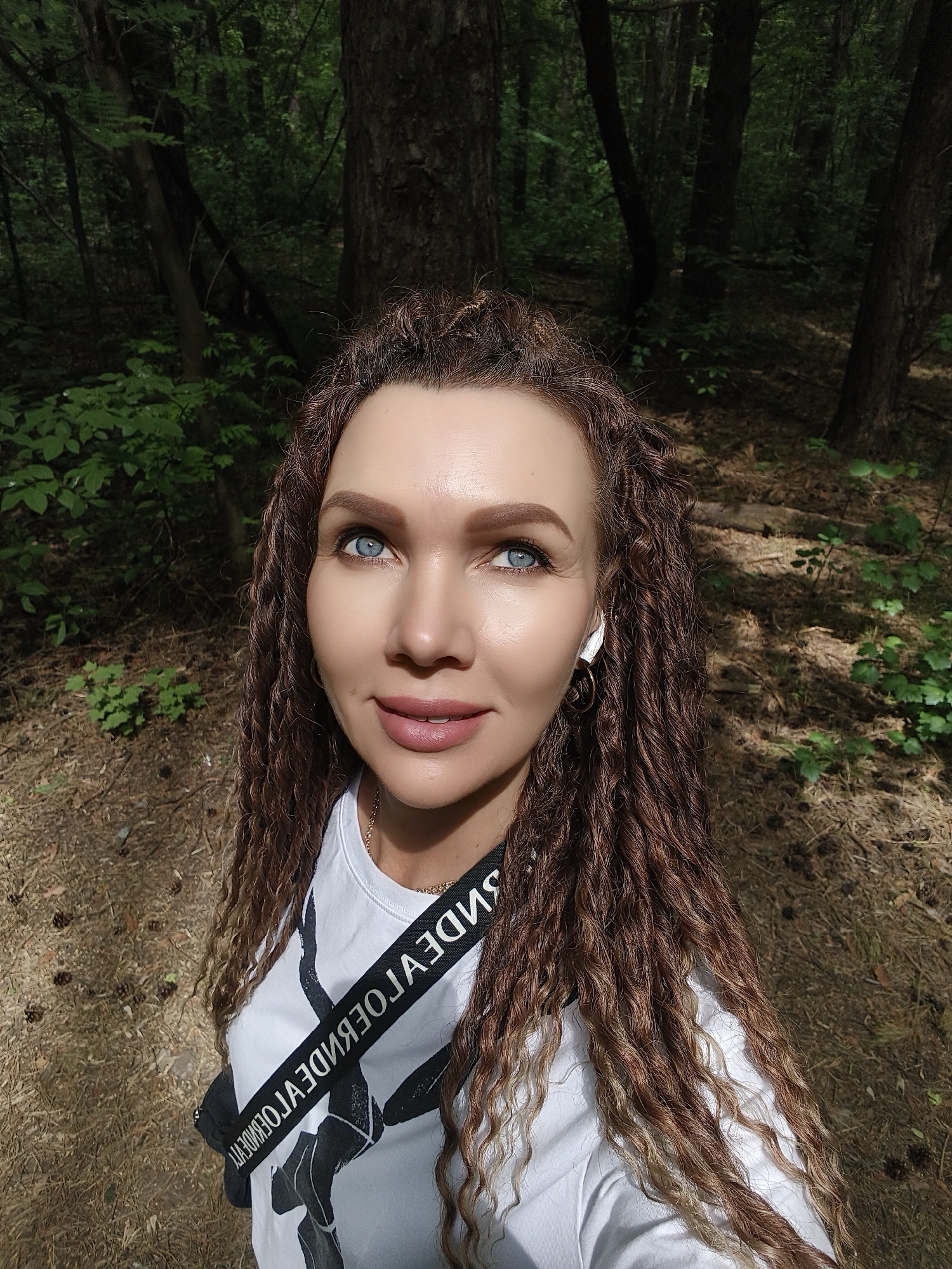 A woman with long, brown, curly hair and striking blue eyes stands in a forest. She is wearing a white t-shirt with a black strap across her torso. She appears to be wearing an earbud in her left ear. The background consists of trees and ground covered with fallen pine needles. The scene is lit with natural sunlight filtering through the trees. The location appears to be a forested area, possibly a park or wooded trail. The city is unknown.