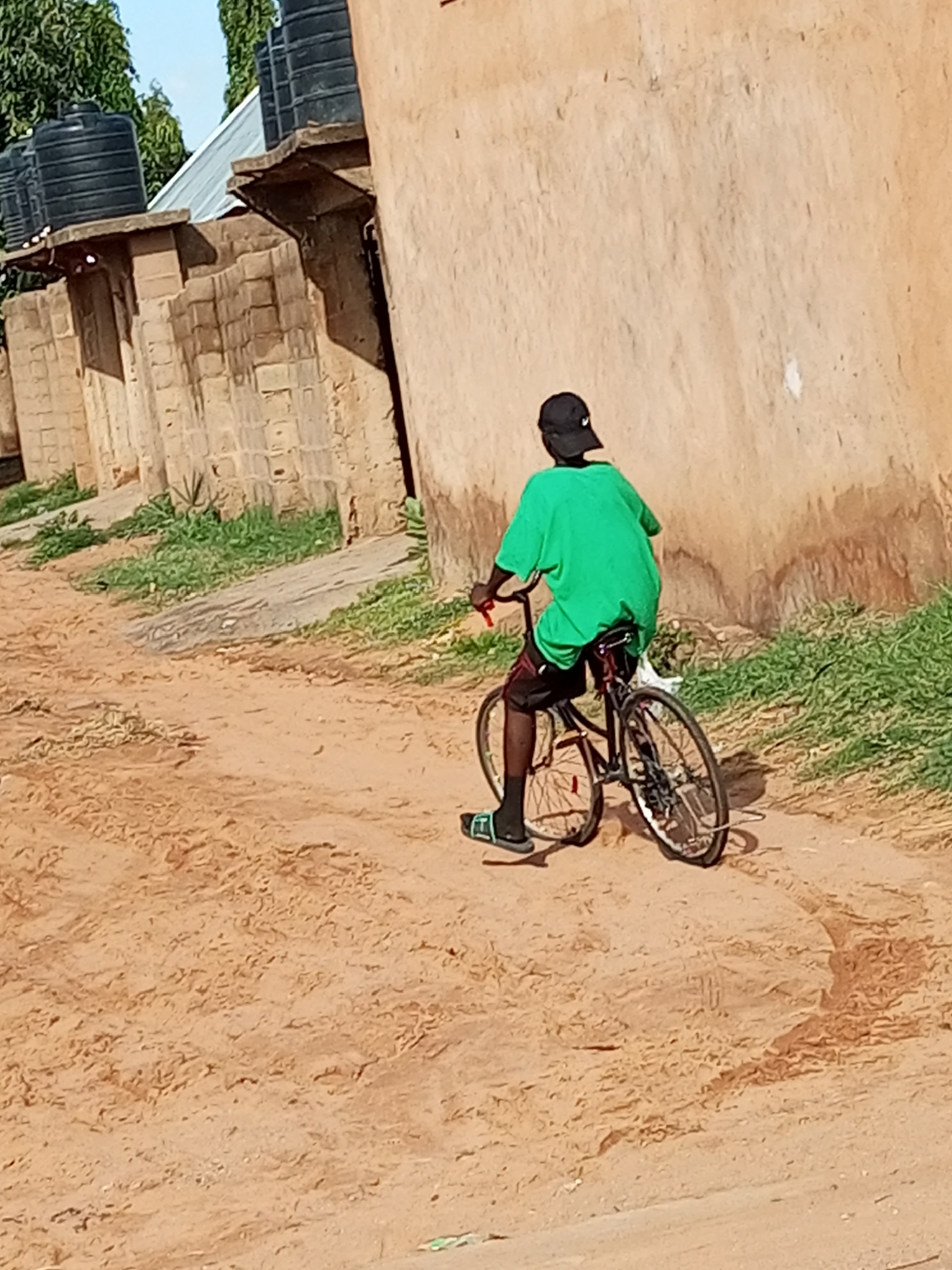 A person wearing a green t-shirt and black shorts rides a bicycle on a dirt path. The person is also wearing a black baseball cap and light-colored sandals. The bicycle has a black frame and two wheels. In the background, there is a light-colored building with a metal roof and several black water tanks. The ground is dry and sandy, with some sparse green vegetation growing along the side of the path. The scene appears to be set in a rural or semi-urban area during the daytime, with bright sunlight casting shadows. The overall impression is one of everyday life in a warm climate. There is no visible text in the image.
