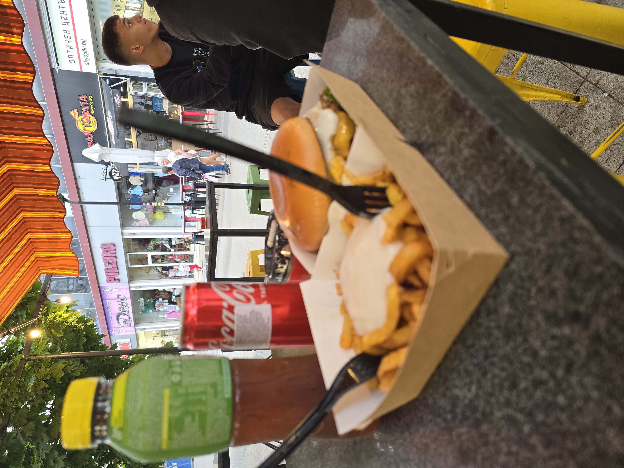 The image captures a close-up, angled view of a meal and beverages on a table in Pleven, Bulgaria. A young man is partially visible in the background. He is wearing a dark-colored long-sleeved shirt and facing away from the primary focus.
The main subject is a burger and fries served in a cardboard container. A black plastic fork is placed inside the container. Next to the meal are a bottle of green juice and a can of Coca-Cola.
In the background, storefronts and pedestrians are visible across the street, one business sign displays the words "КАНТОВАТА express".
The photo appears to have been taken outdoors, at what might be a restaurant or cafe with an orange-and-white awning. A portion of a yellow chair is seen at the top right of the frame.