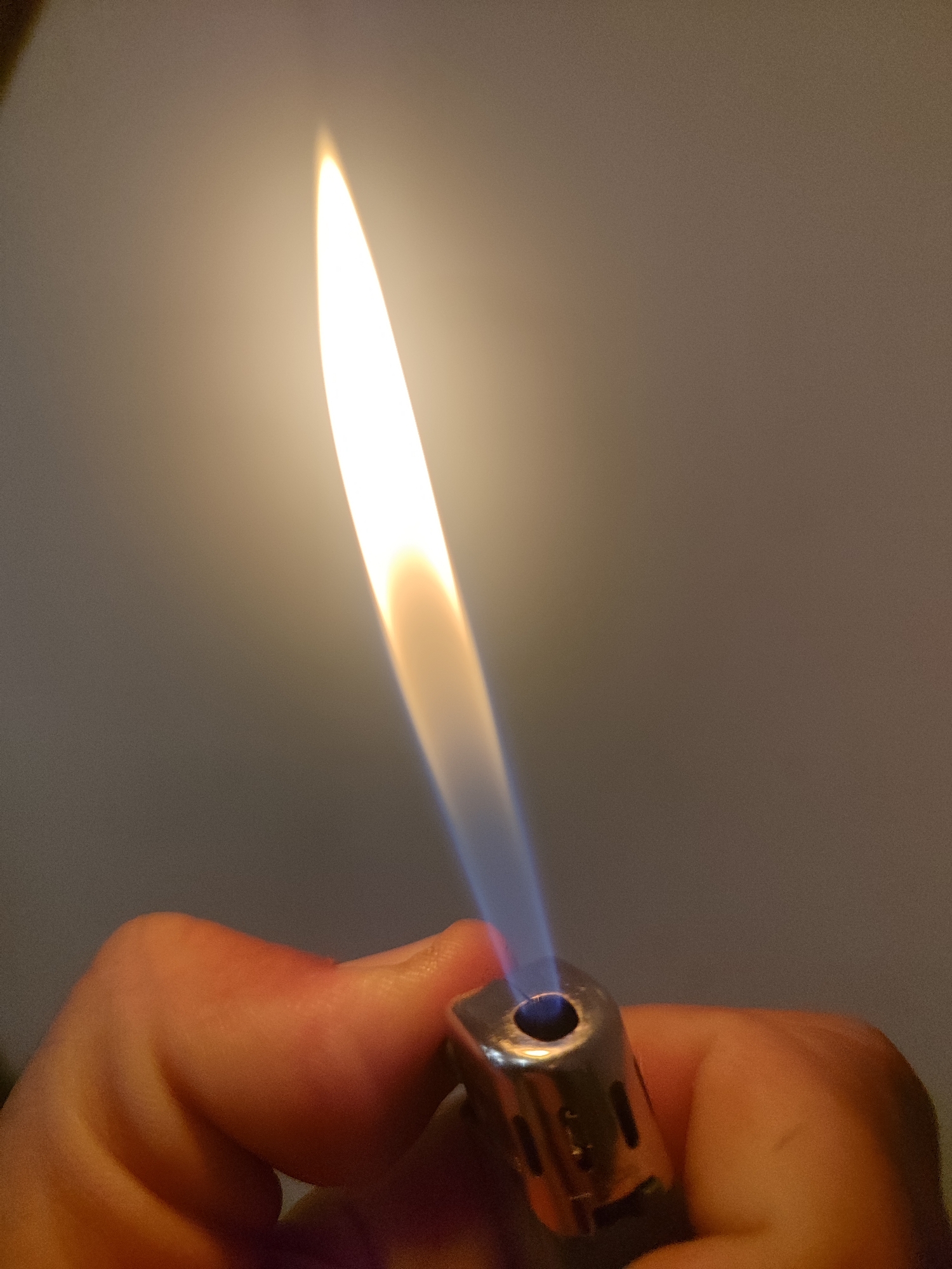The image shows a person holding a lit butane torch lighter. The flame is long, thin, and mostly yellow with a blue base. The lighter is metallic with visible ventilation slots. The person's hand is holding the lighter. The background is a neutral gray. There are no indications of the location being Burgas, Bulgaria.