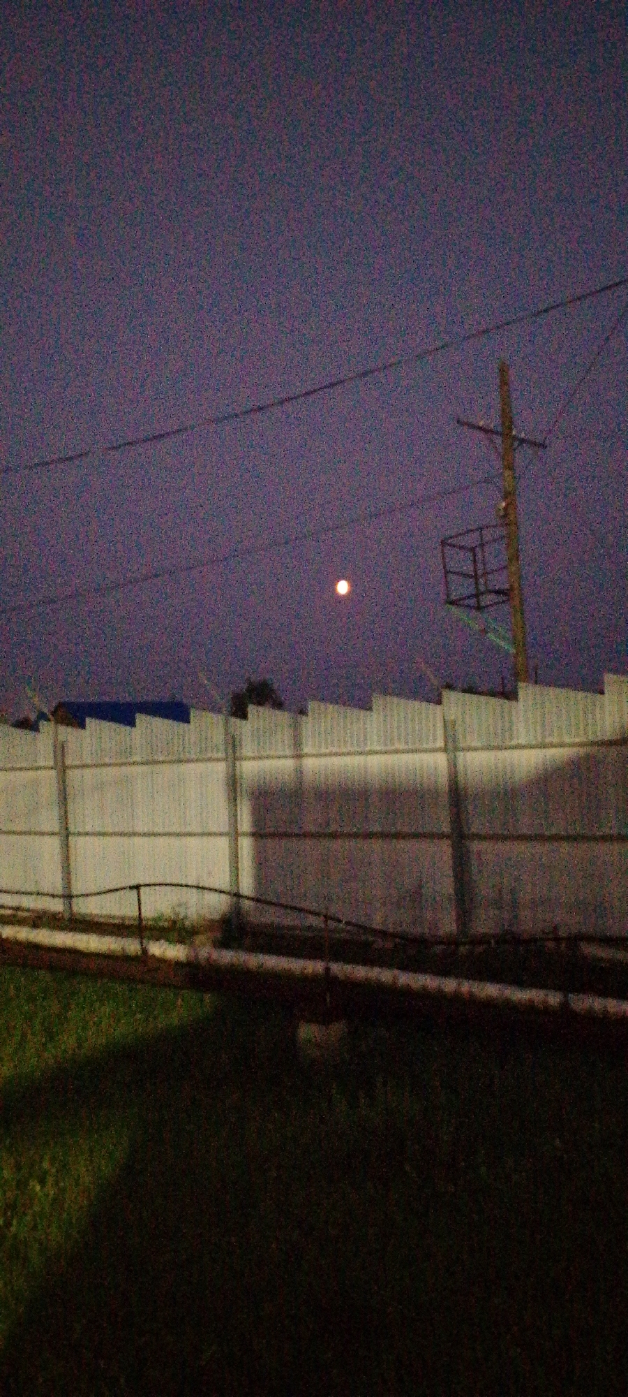 The image depicts an outdoor scene at dusk or night. A tall, wooden utility pole with electrical wires and a small caged platform is visible on the right side of the frame. In the sky, a luminous, round object, likely the moon, is present. In the foreground, a corrugated metal fence runs horizontally across the image, casting a shadow. Below the fence, a dark area of grass is visible. The sky has a purplish hue and is speckled with digital noise, indicating a low-light photograph. The overall impression is one of an industrial or semi-urban environment at twilight. No people or specific landmarks of Tulagino, Russia, are discernible in the image.
