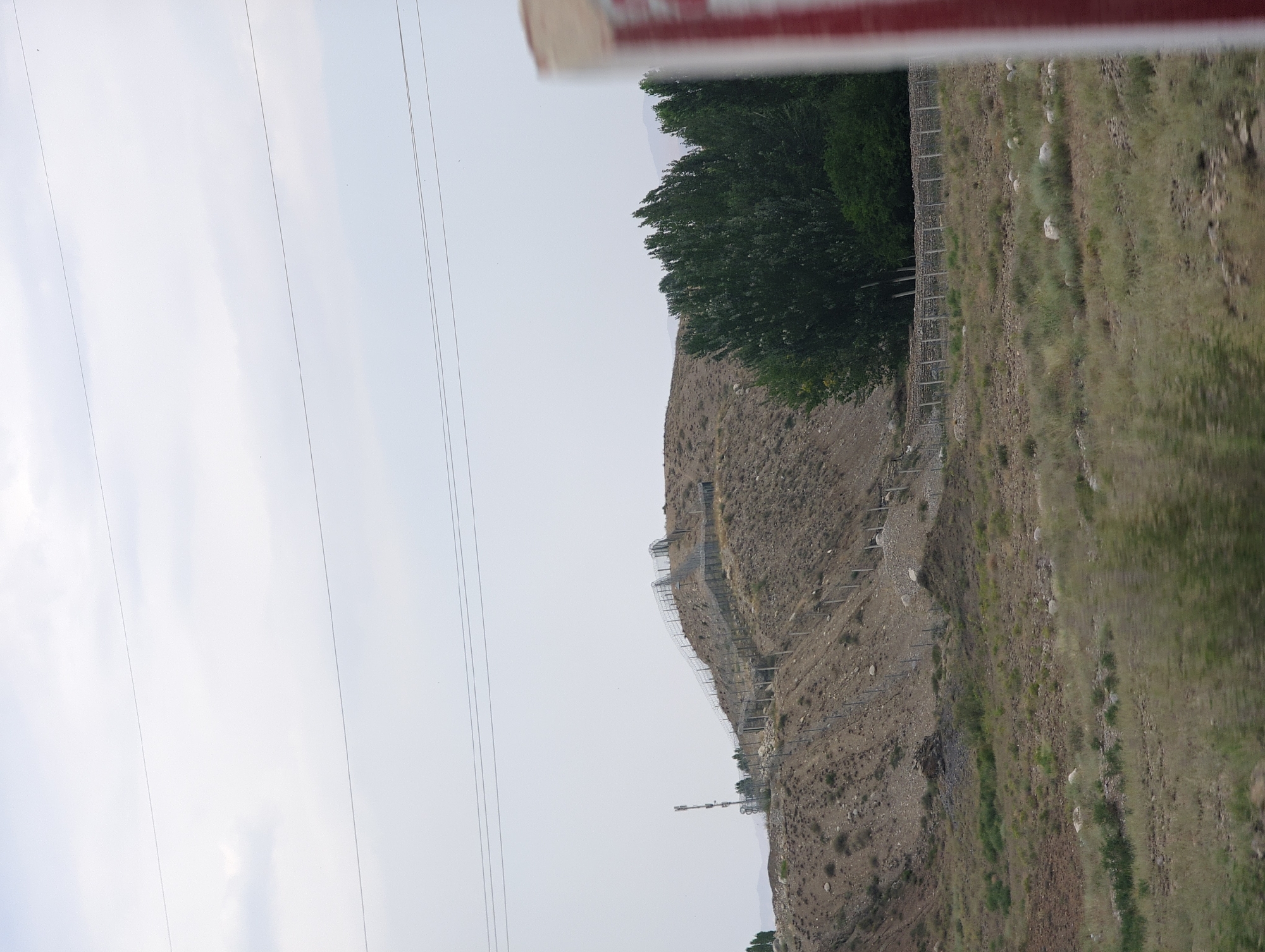 The image shows a fenced hillside with a communication tower on it. The hillside is covered with dry vegetation and rocks. There are trees on the right side of the image. In the foreground, there is a blurred object with red and white stripes, possibly a sign or a vehicle. The sky is overcast. The scene appears to be in a rural or mountainous area, with the provided information suggesting the location is Көк-Таш, Kyrgyzstan. The fencing appears to be a security measure, possibly a border fence or a fence around a protected area. The communication tower suggests the presence of infrastructure in the area.