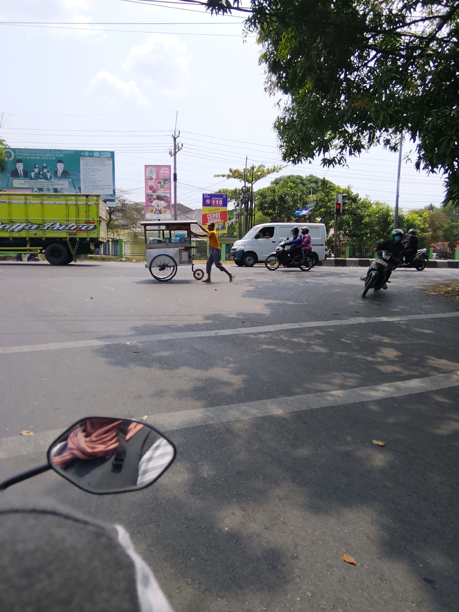 The image shows a street scene in Cirebon, Indonesia. A yellow truck is parked on the left side of the road, and a person is pushing a food cart in the middle of the street. Several motorcycles are also visible, with riders wearing helmets. A white van is parked on the right side of the road, and traffic lights are visible in the background. The sky is clear and sunny, with some clouds. Trees and buildings can be seen on the sides of the road. The scene appears to be a busy street in a commercial area.