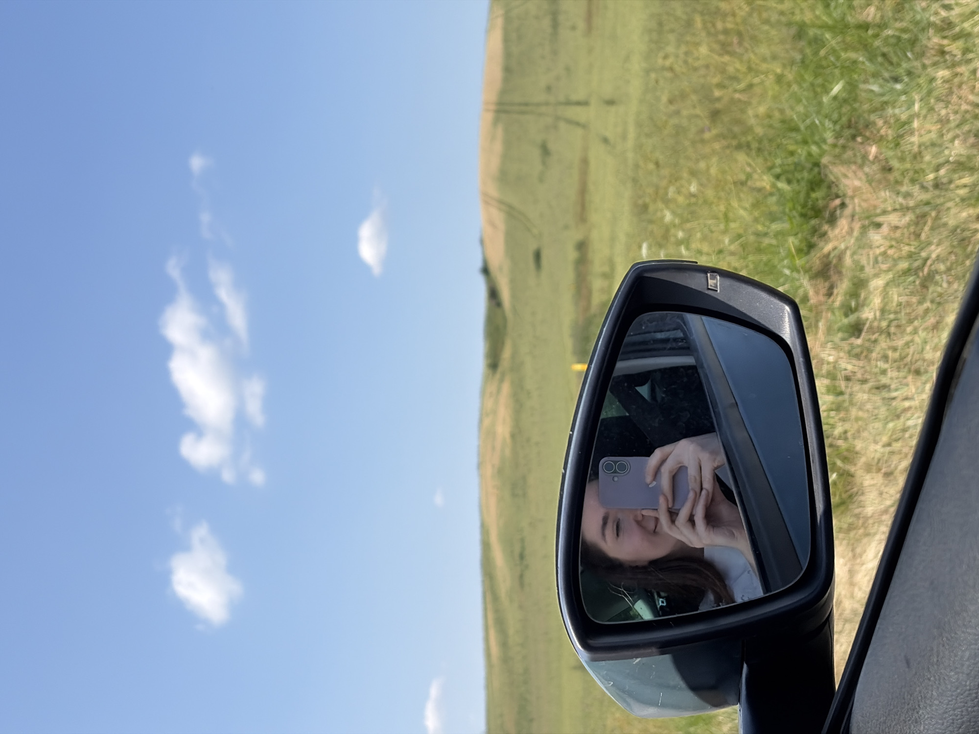 In the foreground, a car's side mirror reflects a young woman holding a purple phone and taking a selfie. Her eyes are partially closed, and she is smiling. The background of the mirror shows the interior of a car. Outside the mirror, a vast, grassy field stretches towards rolling hills under a clear blue sky with scattered white clouds. The field has visible tracks, suggesting agricultural activity. The scene is set in the countryside, possibly in the Astrashitskagaradokski rural council, Belarus, given the landscape.