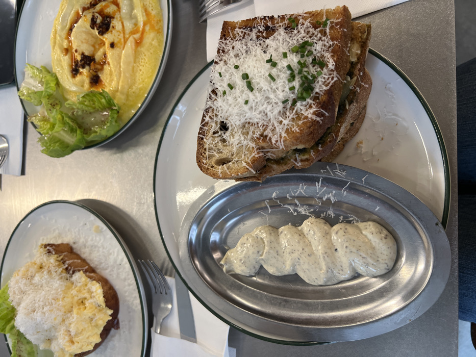 The image displays an overhead view of a dining table in an indoor setting, identified as Paris, France, set for a meal. Three white plates with green rims are arranged on a light grey table surface. The upper left plate contains a slice of toasted bread topped with scrambled eggs and covered with a dense layer of grated white cheese. The upper right plate holds a creamy yellow dish, possibly hummus or polenta, garnished with a central swirl of reddish-orange chili oil and dark specks, alongside several small green lettuce leaves. In the foreground, a grilled sandwich, likely a croque monsieur, made with toasted bread, is presented on a plate. It is heavily topped with grated white cheese and sprinkled with chopped green chives. Adjacent to the sandwich, a small, oval, metallic dish contains a neatly piped, light-beige, textured creamy sauce with dark specks. Silverware, specifically forks, and white paper napkins are visible on the table. The lower edge of the frame reveals the top of a person's legs in blue jeans, indicating a diner is seated.