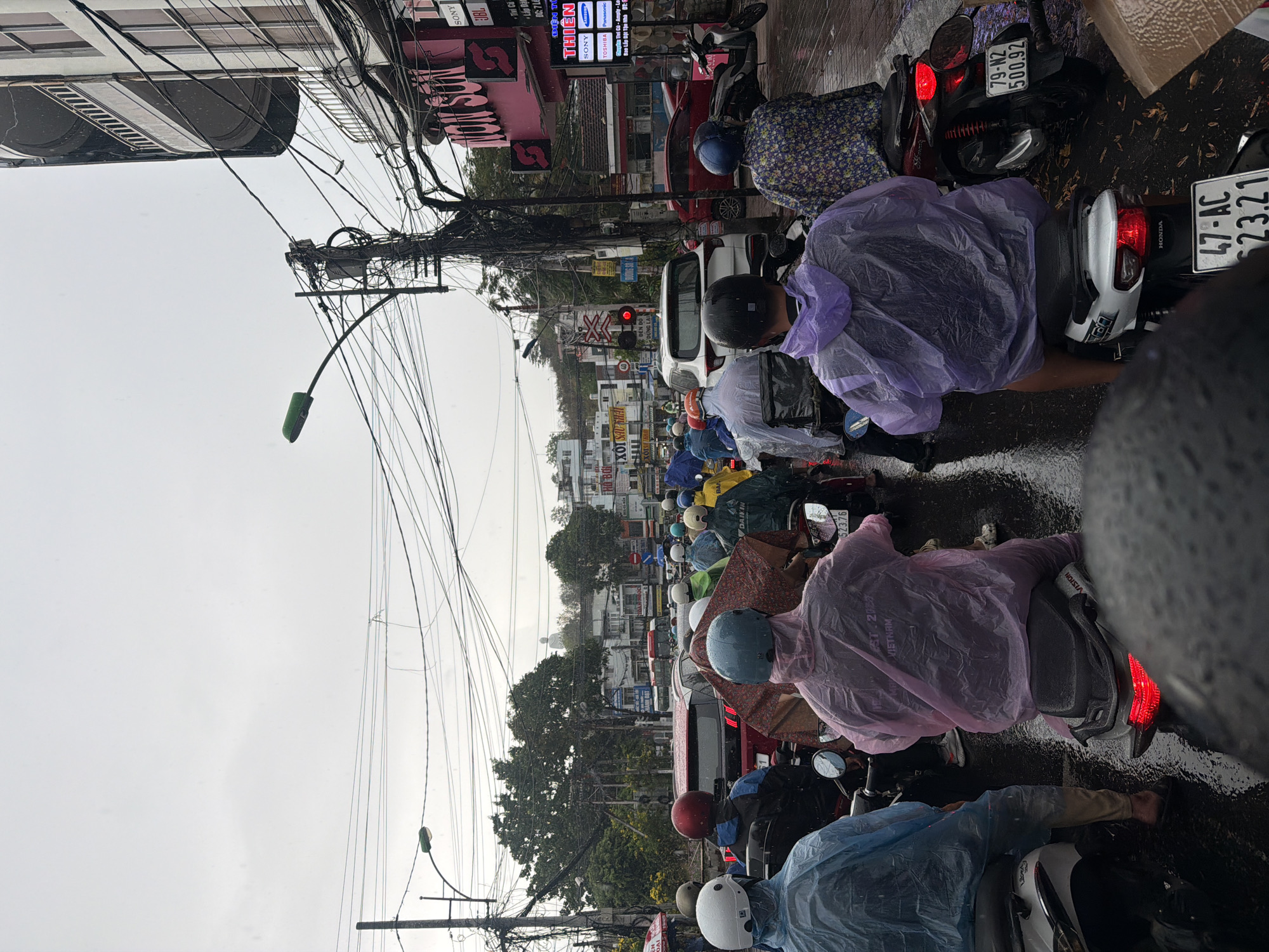 The image captures a street scene in Nha Trang Ward, Vietnam, under what appears to be rainy weather. Many people on motorbikes, most wearing rain ponchos, are lined up on the wet road. The vehicles are densely packed, suggesting heavy traffic. Overhead, a complex network of tangled power lines is visible against the overcast sky. Buildings and shops line the street, with signs for brands like "SONY," "TOSHIBA," and "DAIKIN." Motorcycle license plates are visible, including "47-AC 23.21" and "79-N2 500.92." A red traffic light is also visible.