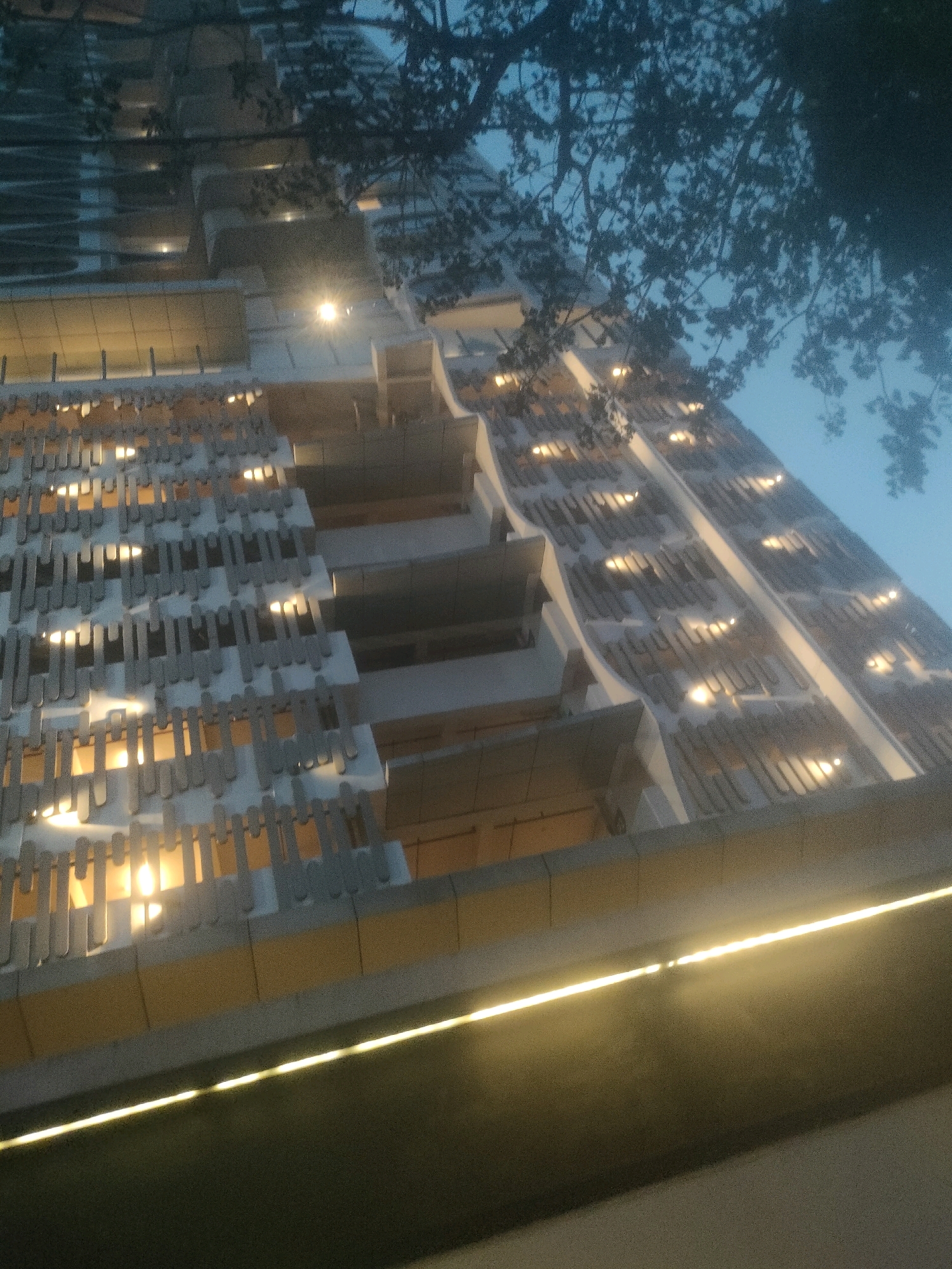 This is a low-angle, nighttime exterior shot of a modern, multi-story building in Surabaya, Indonesia. The building features a unique facade with alternating panels of light-colored, decorative elements and darker openings, behind which warm light is visible, suggesting interior illumination. The building's design includes curved sections and protruding balconies or ledges. A horizontal line of bright yellow light runs along the bottom edge of the frame, possibly an exterior lighting feature of the building or a separate structure. Silhouetted tree branches with leaves are visible in the upper right portion of the image against a dark blue sky. The overall impression is one of urban architecture at night, with architectural lighting playing a significant role in the visual aesthetic.