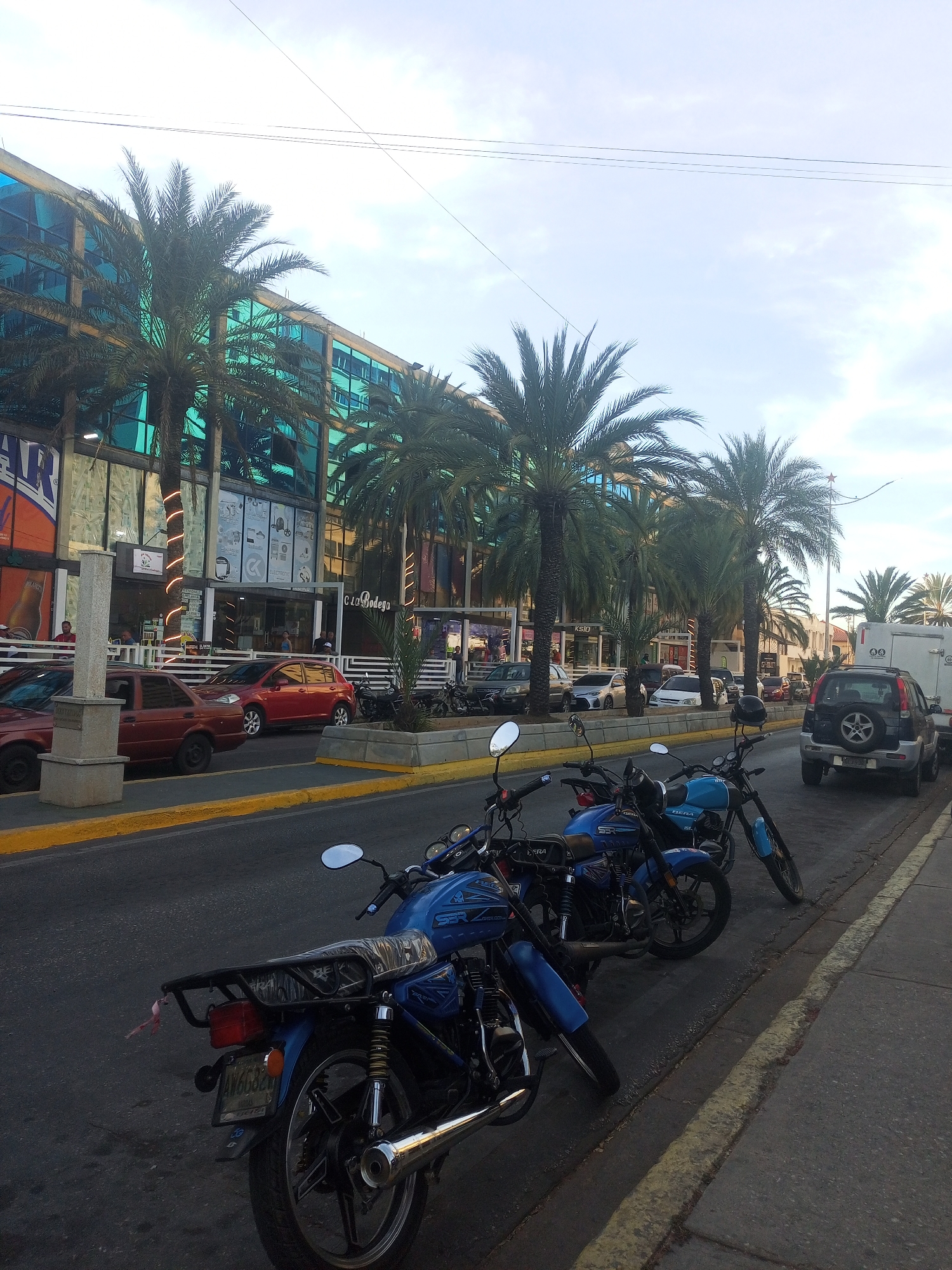 The image shows a street scene in Juan Griego, Venezuela. Several blue motorcycles are parked along the right side of the road. A gray SUV is parked further down the street. Cars are driving on the road. On the left side of the street are buildings with turquoise-tinted windows and palm trees in front. Some people are standing and sitting outside of the buildings.