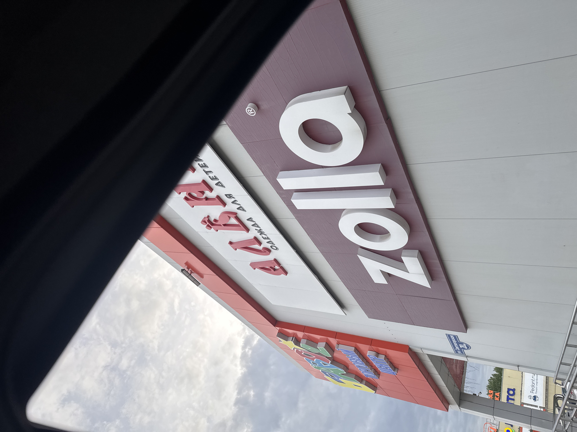 The image captures an upward view of the facades of several stores in Perm, Russia, focusing on their signage. A Zolla store sign, rendered in white block letters against a maroon and white background, is centrally featured. Adjacent to it, the Алби (Albi) store sign, written in Russian, also stands out with its red lettering indicating "Clothes for Children." The lower portion of the frame displays a colorful section of another storefront with letters that are partially visible. A street sign "ул. Светлогорская 20" is seen. In the background, additional storefront signs, including one for "Pedant.ru" (a smartphone repair service), are visible. The sky, visible between the buildings, is overcast. The photograph is taken from an unusual angle, likely from inside a vehicle, as a dark portion of the car's interior is visible on the left side of the frame.
