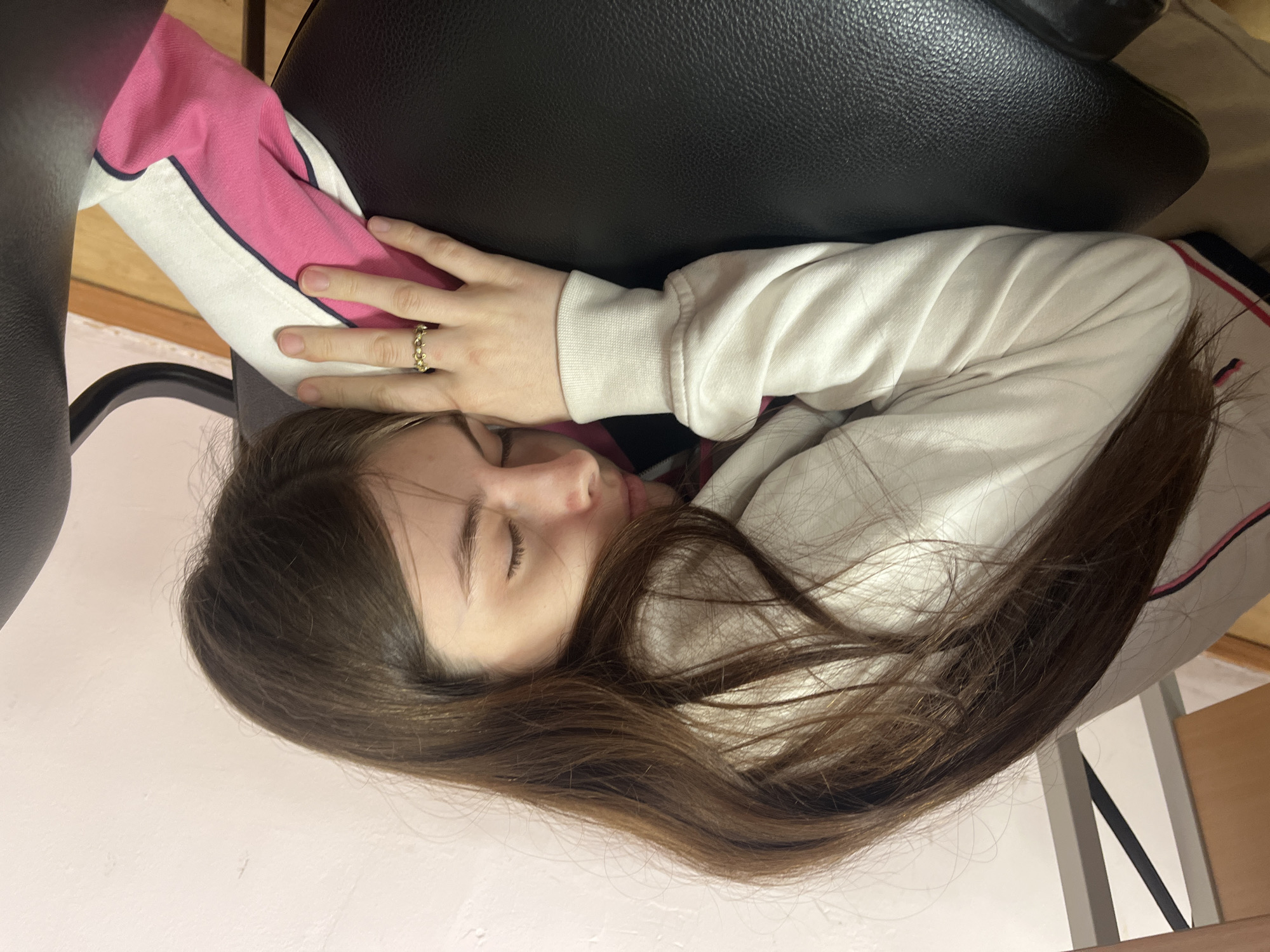 A young woman with long brown hair is sleeping on a black office chair. She is wearing a white sweatshirt with pink and dark blue stripes on the sleeves and a ring on her finger. The scene appears to be indoors, possibly in a home or office. No information can be identified about the location in the city of Saint Petersburg, Russia.