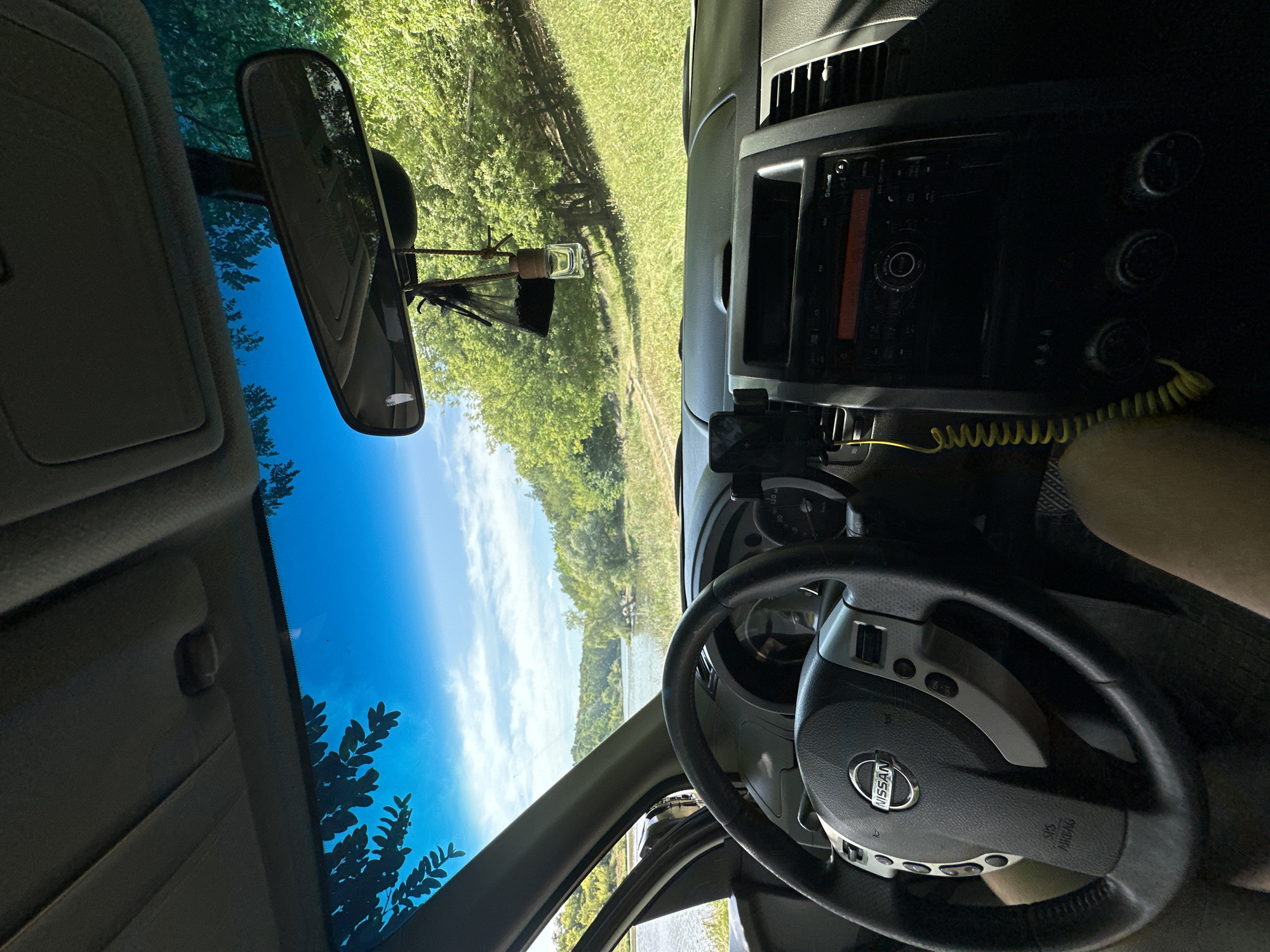 An interior shot of a Nissan car shows the dashboard and steering wheel in the foreground, with a view of a lake and a wooded hillside through the windshield. A cell phone is mounted on the dashboard, and an air freshener hangs from the rearview mirror. The scene appears to be a scenic overlook, possibly near the city of Makiv, Ukraine, suggesting a moment of peaceful contemplation or rest during a drive.