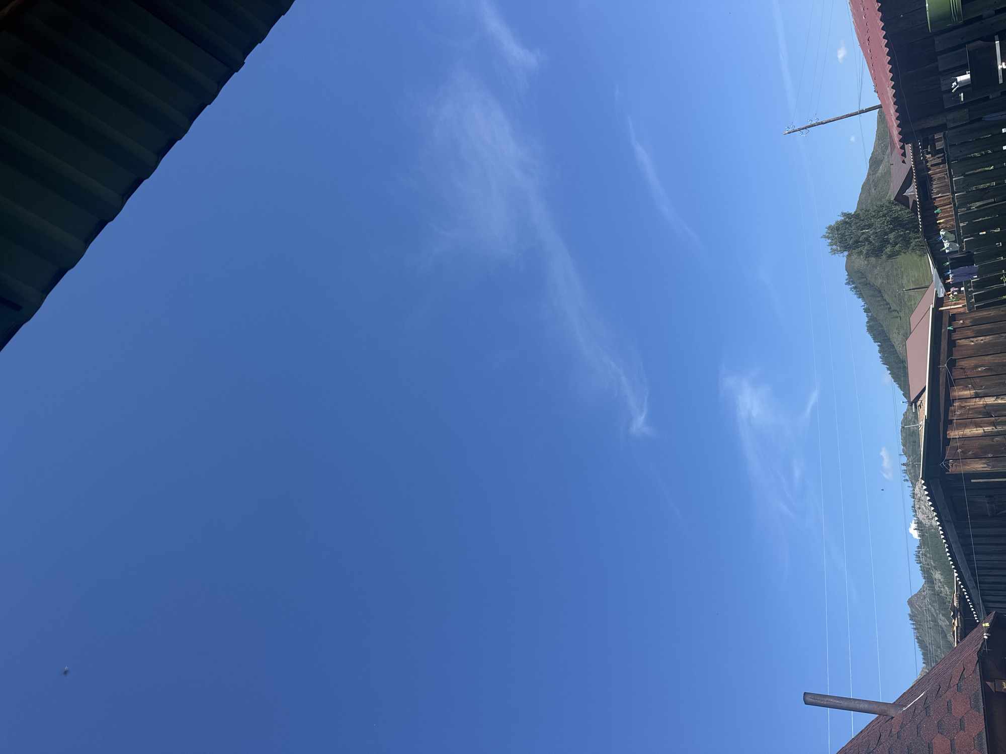 The image depicts a view of the sky and rooftops in Yabogan, Russia. The sky is a clear, bright blue with sparse, wispy white clouds. In the foreground, several rooftops are visible, including a dark green corrugated metal roof partially obscuring the top right of the frame. Below this, various wooden structures with corrugated and shingled roofs are present, suggesting a residential or semi-rural area. Laundry hangs on a line between two wooden structures. In the background, a rolling, tree-covered mountain range is visible under the expansive sky. The overall impression is that of a sunny day in a mountainous region.