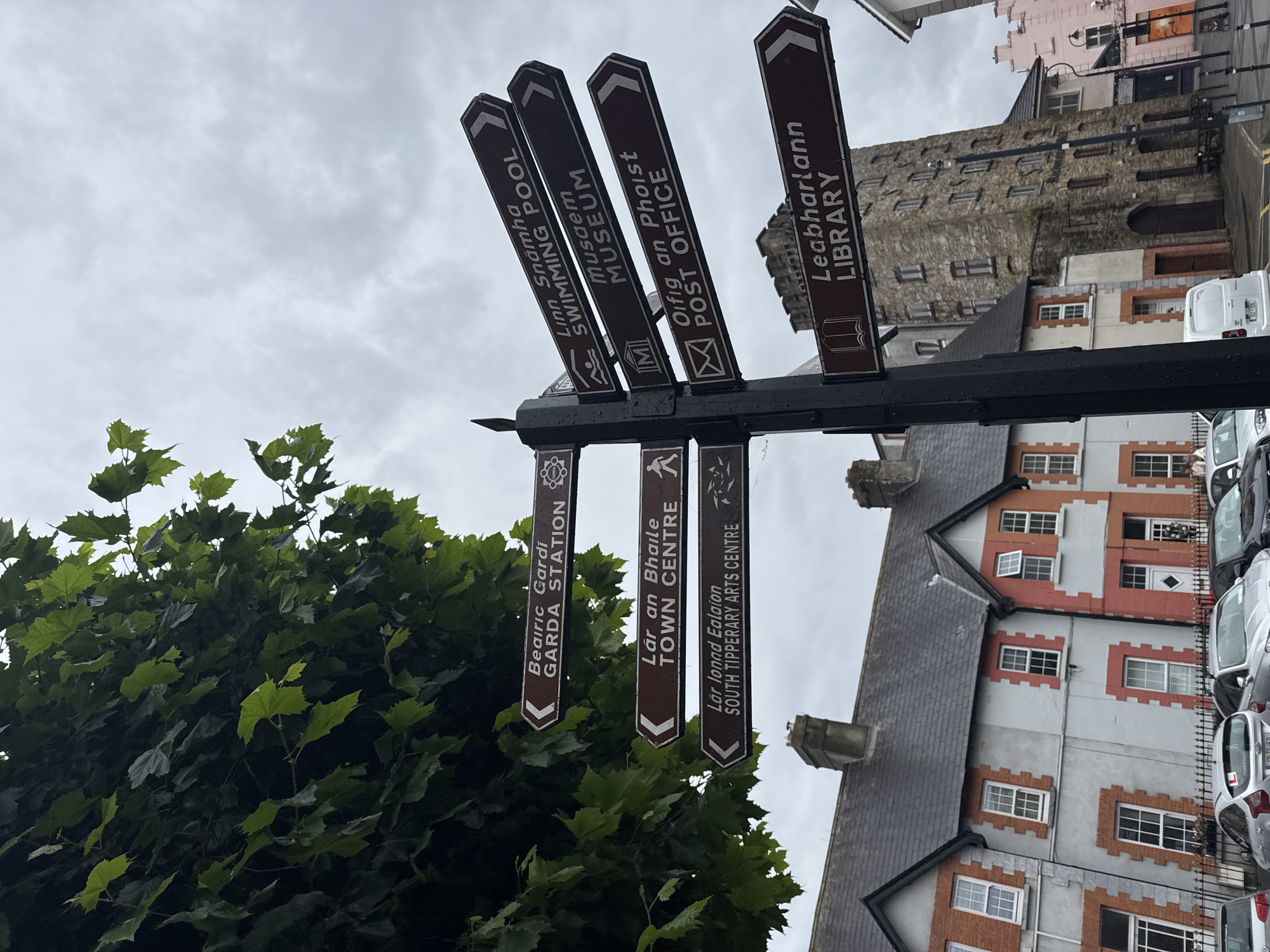 In Clonmel, Ireland, a multi-directional signpost stands prominently on a street. The signpost, made of dark metal, displays several brown directional signs with white lettering. These signs point to various locations: "Beairic Gardi GARDA STATION," "Lár an Bhaile TOWN CENTRE," "Lár Ionad Ealaíon SOUTH TIPPERARY ARTS CENTRE," "Linn Snámha SWIMMING POOL," "Musaem MUSEUM," "Oifig an Phoist POST OFFICE," and "Leabharlann LIBRARY." Some signs include small pictograms representing their destinations, such as a police badge, a person walking, a building, a swimmer, a museum artifact, a letter, and a book.
Behind the signpost, a dense green tree with large leaves partially obscures the sky. The sky itself is overcast and gray. In the background, several buildings are visible. To the left, a row of traditional terraced houses with red brick accents and gray roofs lines the street. To the right, a distinctive stone tower with many windows, resembling an old building or castle, stands tall. Other buildings with varying architectural styles and colors, including a pink facade, are also present. The street appears to be paved, and several parked cars are visible in the foreground and along the side of the road. The overall scene depicts a street in a town, with clear directional information pointing towards local amenities and points of interest.