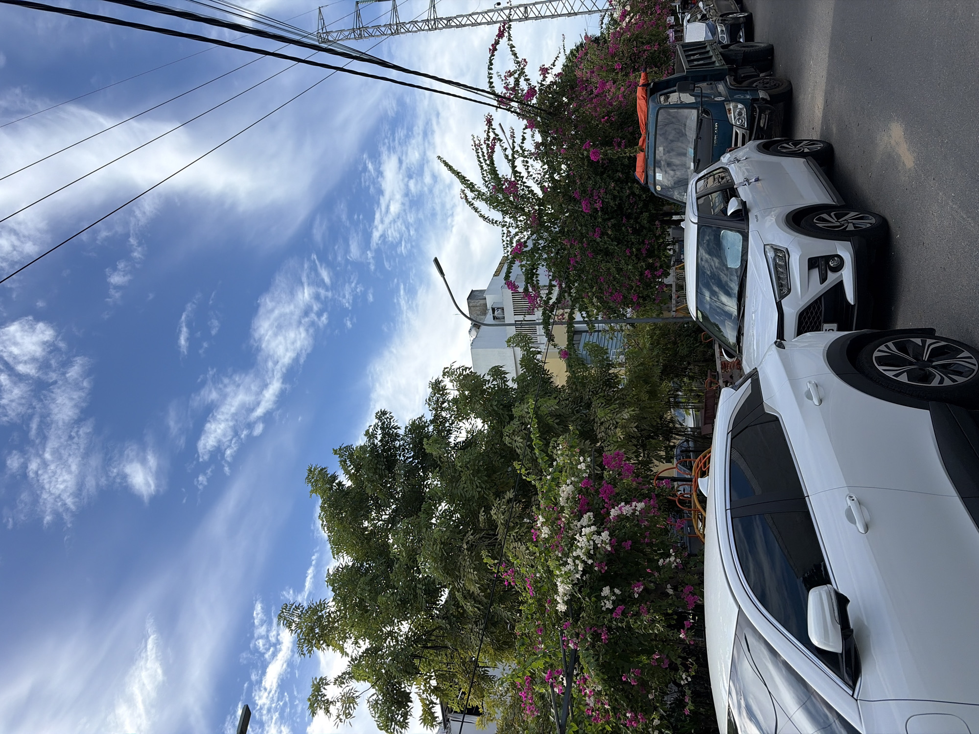 The image captures a street scene in Da Nang, Vietnam, under a bright blue sky with scattered wispy clouds. A row of vehicles is parked along the street. In the foreground, the rear portion of a white SUV is visible on the left. Next to it, a white compact SUV is parked, facing away from the camera. Further down the street, a dark green dump truck with an orange tarp covering its bed is parked, followed by other cars.
The street is lined with lush greenery and flowering bushes, with vibrant pink and white bougainvillea flowers cascading over a trellis structure. In the background, a multi-story building with white and yellow facades is visible, along with a street lamp. Overhead, power lines are strung across the sky, supported by a tall transmission tower on the right side of the image, indicating an urban or suburban setting. The overall scene depicts a typical day in a Vietnamese city, with parked vehicles and abundant vegetation.