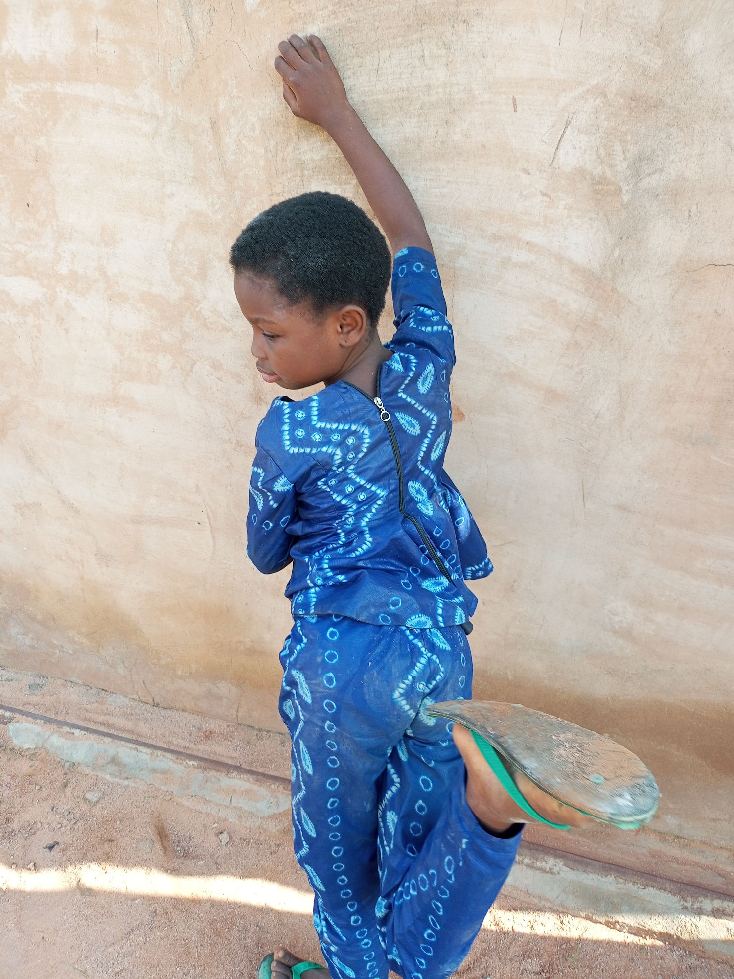 In the city of Tempure, Nigeria, a young child with dark skin and black hair is leaning against a textured, light brown wall. The child is wearing a bright blue patterned outfit with a zipper closure. Their right arm is extended upwards and bent, with their hand resting on the wall. The child's left leg is bent and extended outwards, and they are wearing a sandal on their left foot. The ground beneath the child appears to be dusty and uneven, with some shadows cast on it.
